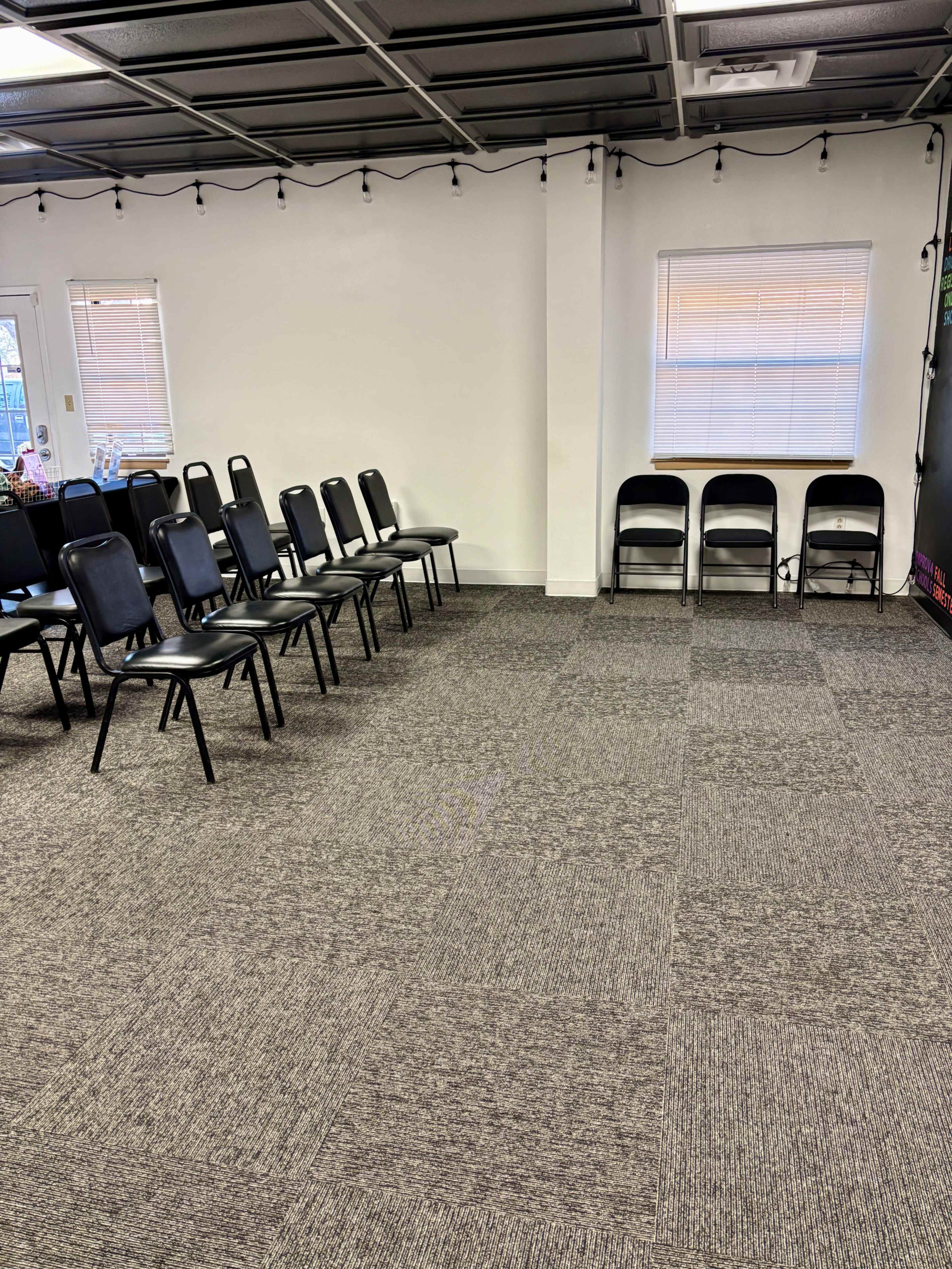 The image shows a room with a section of black chairs arranged in rows on a carpeted floor, alongside two empty folding chairs against the wall.