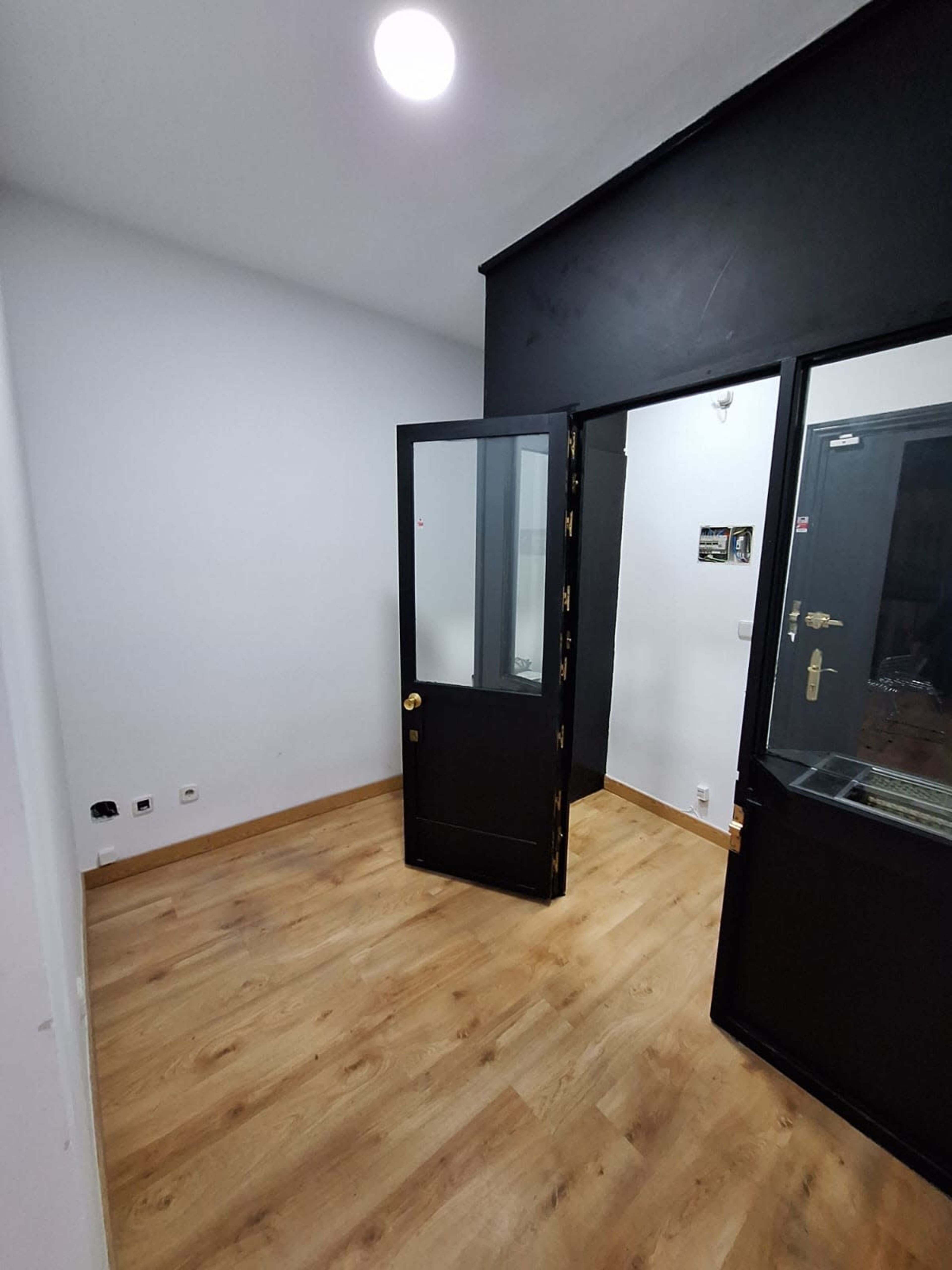 The image shows a small room with light-colored walls, a wooden floor, and a black door that is partially open.