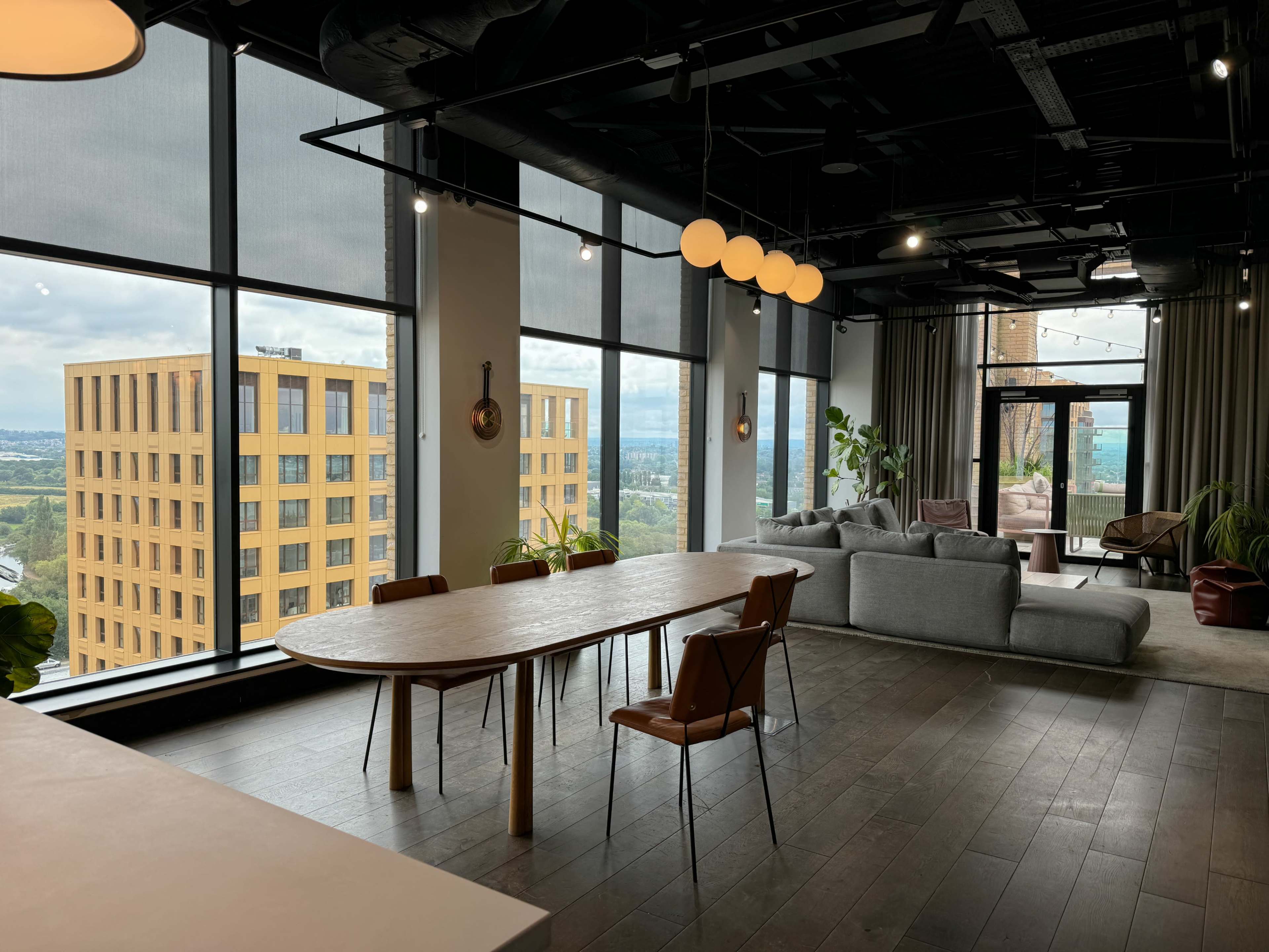 A modern, spacious room features a large table and chairs, with floor-to-ceiling windows that provide a view of the city skyline and surrounding landscape.