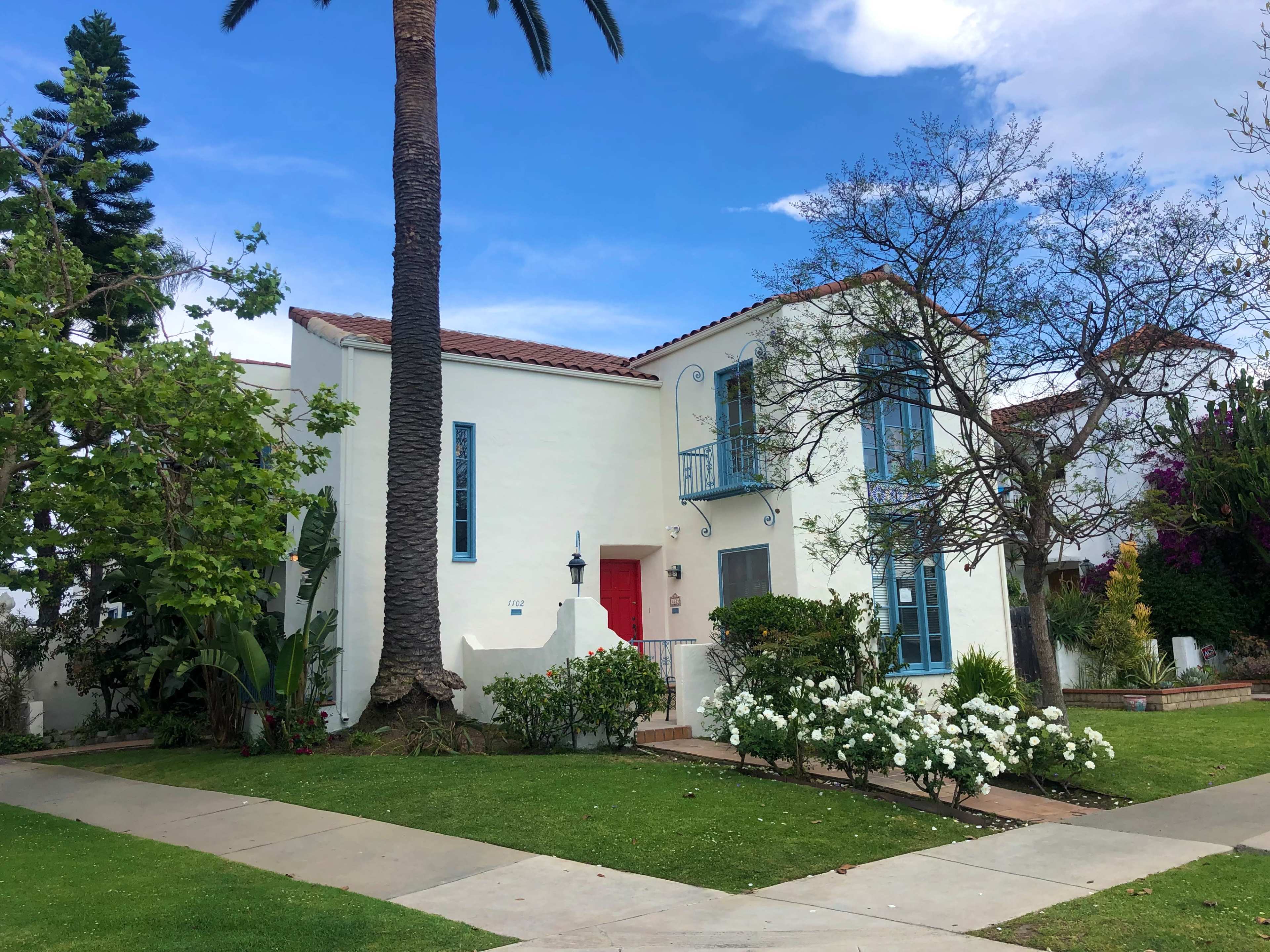A two-story white house with blue-framed windows and a red door is surrounded by greenery and flowers.