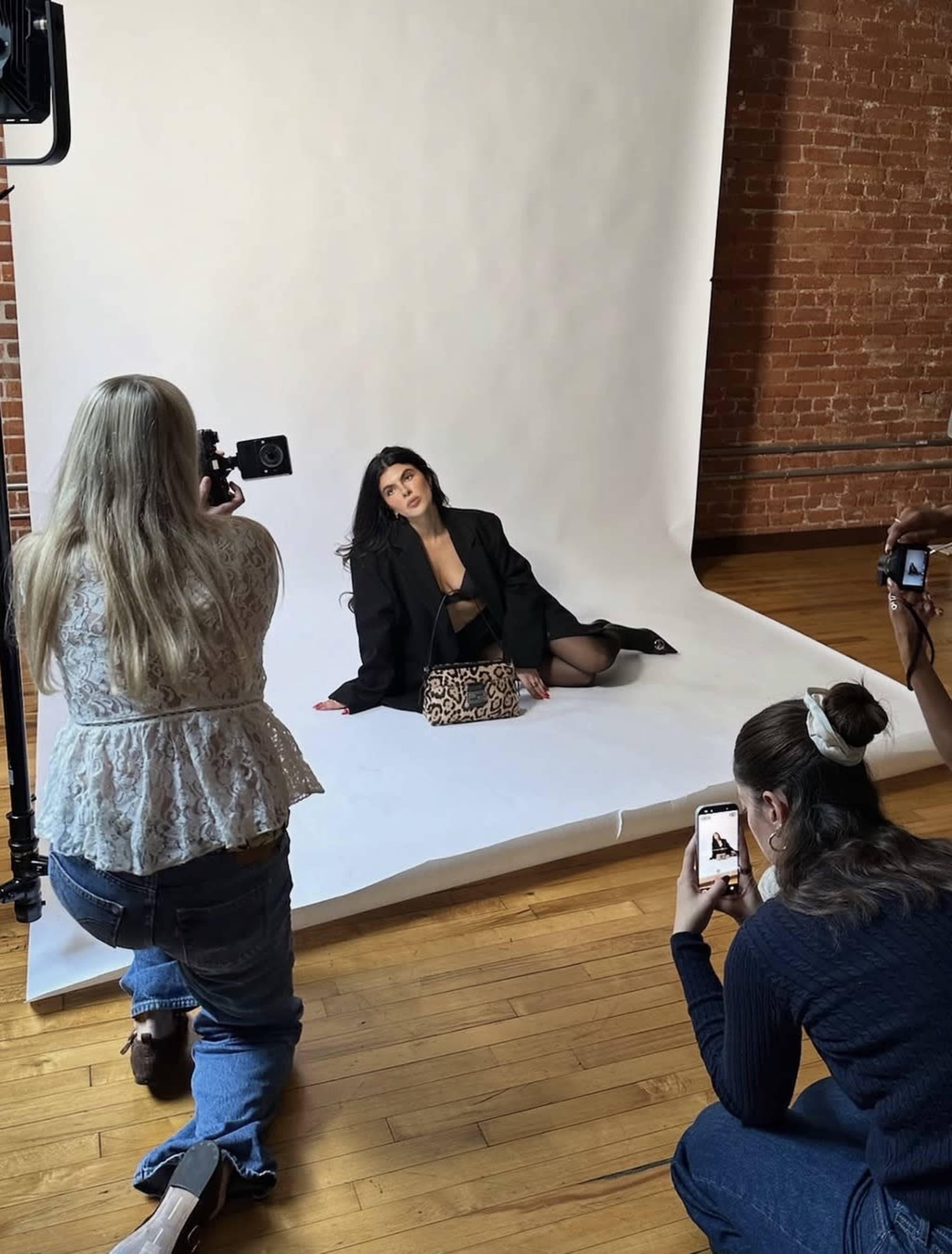 In a photo studio with a brick wall backdrop, a model poses with a handbag while two photographers capture her from different angles.
