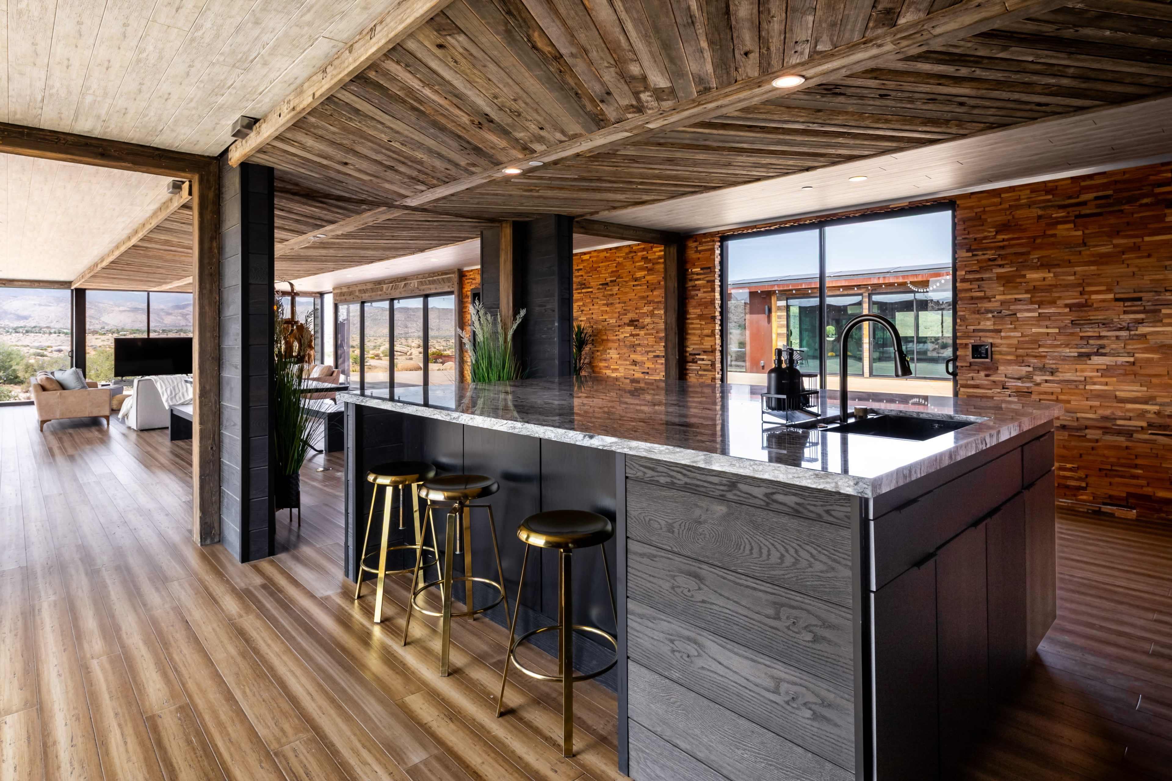 The image shows a modern kitchen with a large island, sleek countertops, and bar stools, surrounded by large windows that provide a view of the outdoors.