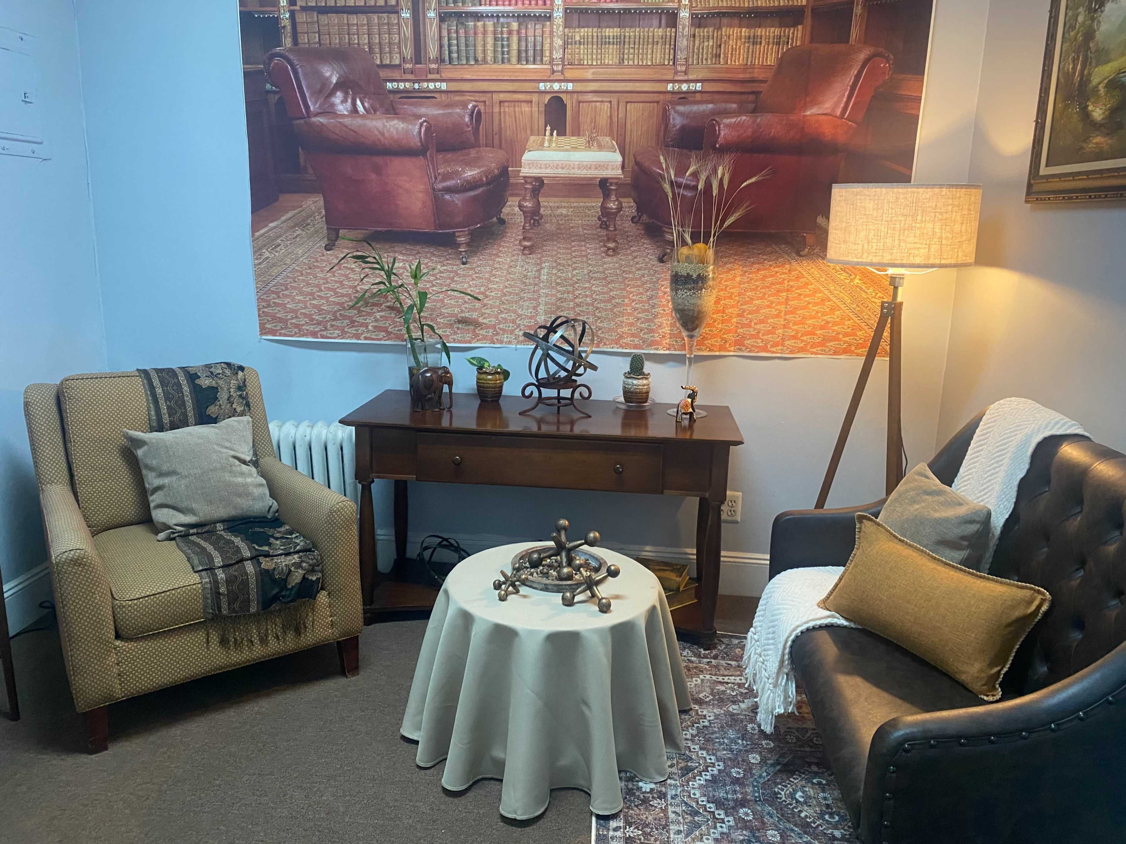 A cozy sitting area with two armchairs, a small table adorned with decorative items, and a backdrop of a library-like setting.