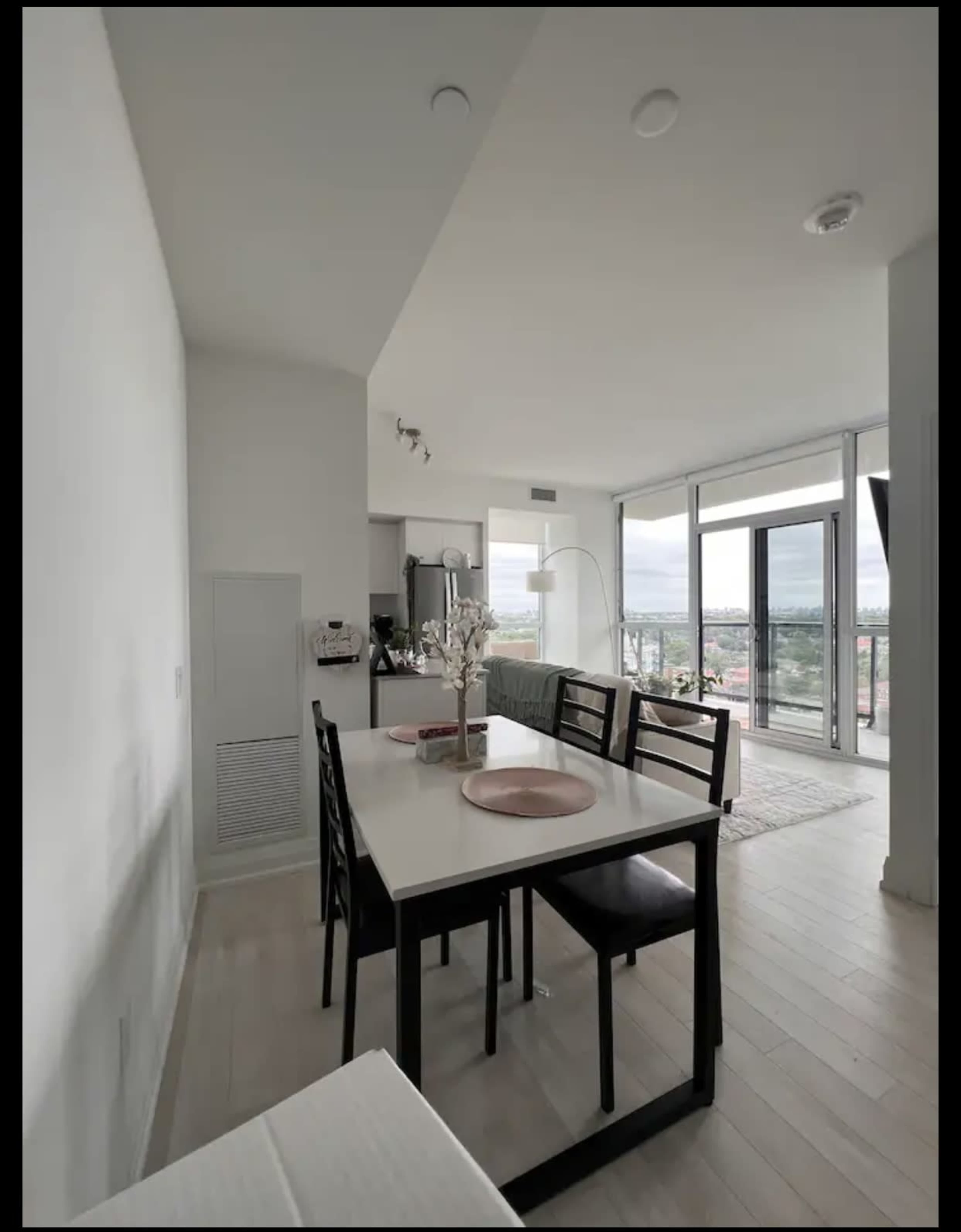 A modern dining area features a rectangular table with black chairs, leading to a spacious living area with floor-to-ceiling windows and a view of the outside.