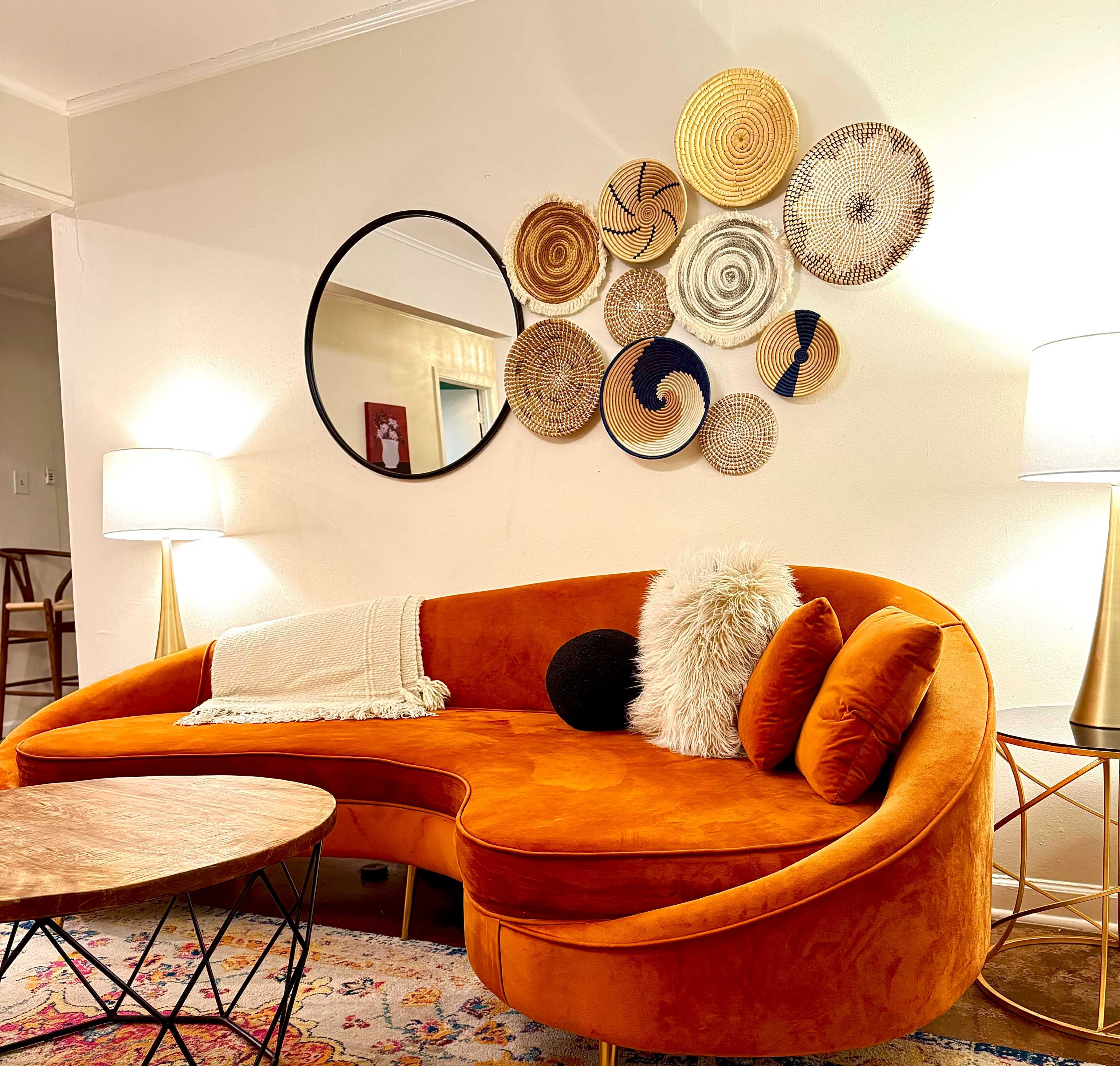 A modern living room featuring an orange sectional sofa with decorative throw pillows, a round mirror, and an arrangement of woven wall art above the couch.