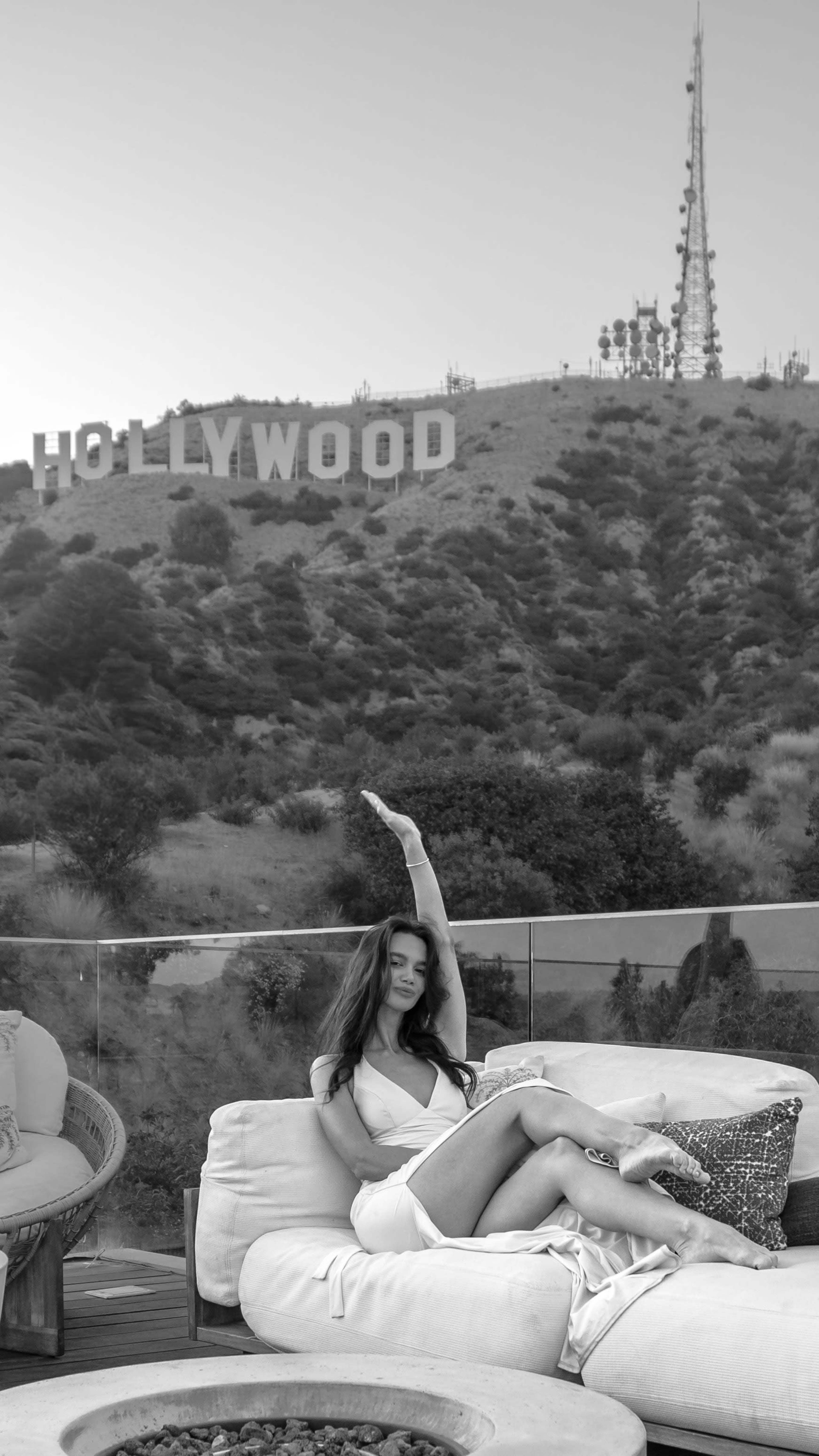 A woman sits on a couch with her arm raised, while the Hollywood sign is visible on a hillside in the background.