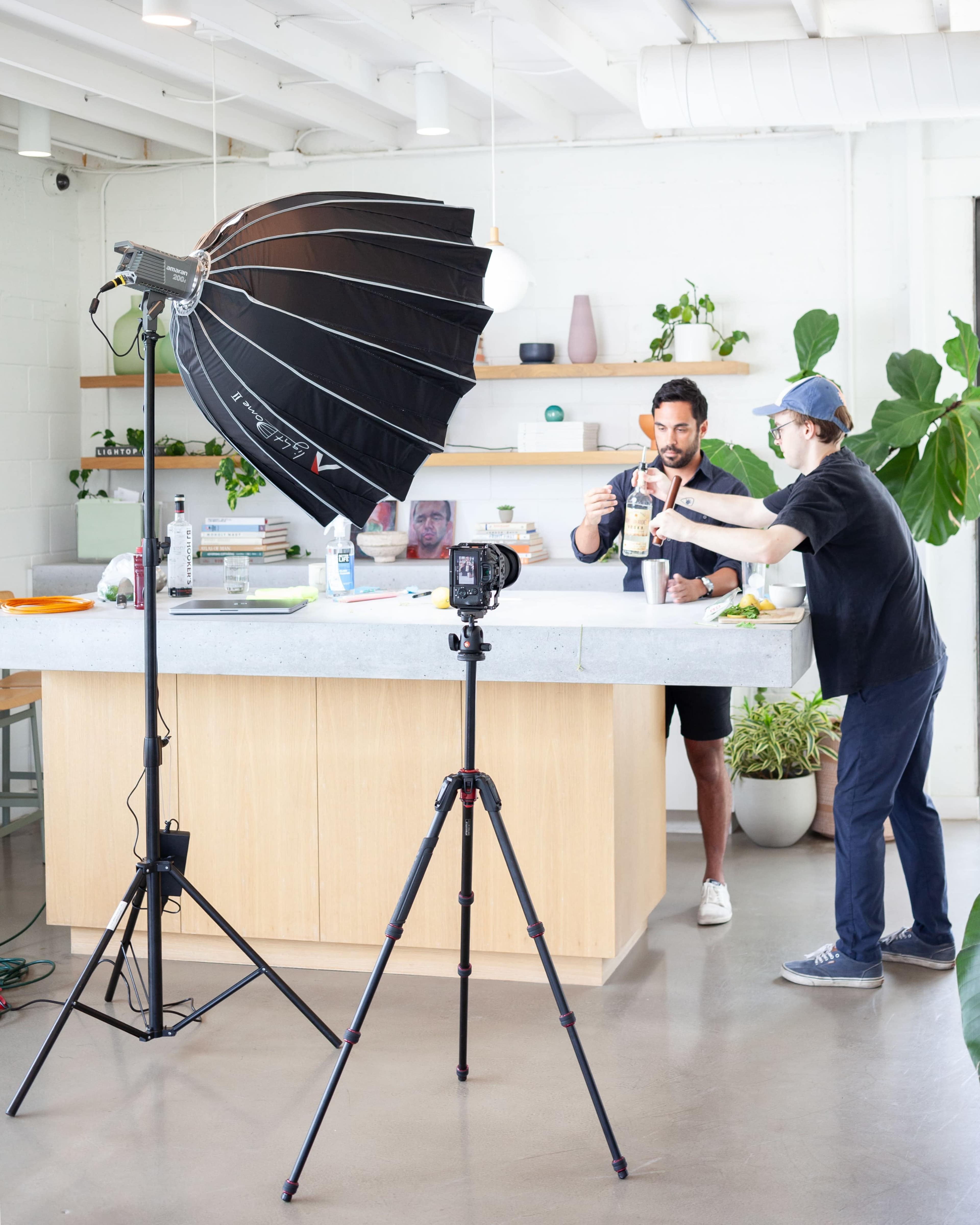 A man prepares a drink at a kitchen counter while a person assists him, with a large softbox light and camera set up nearby.