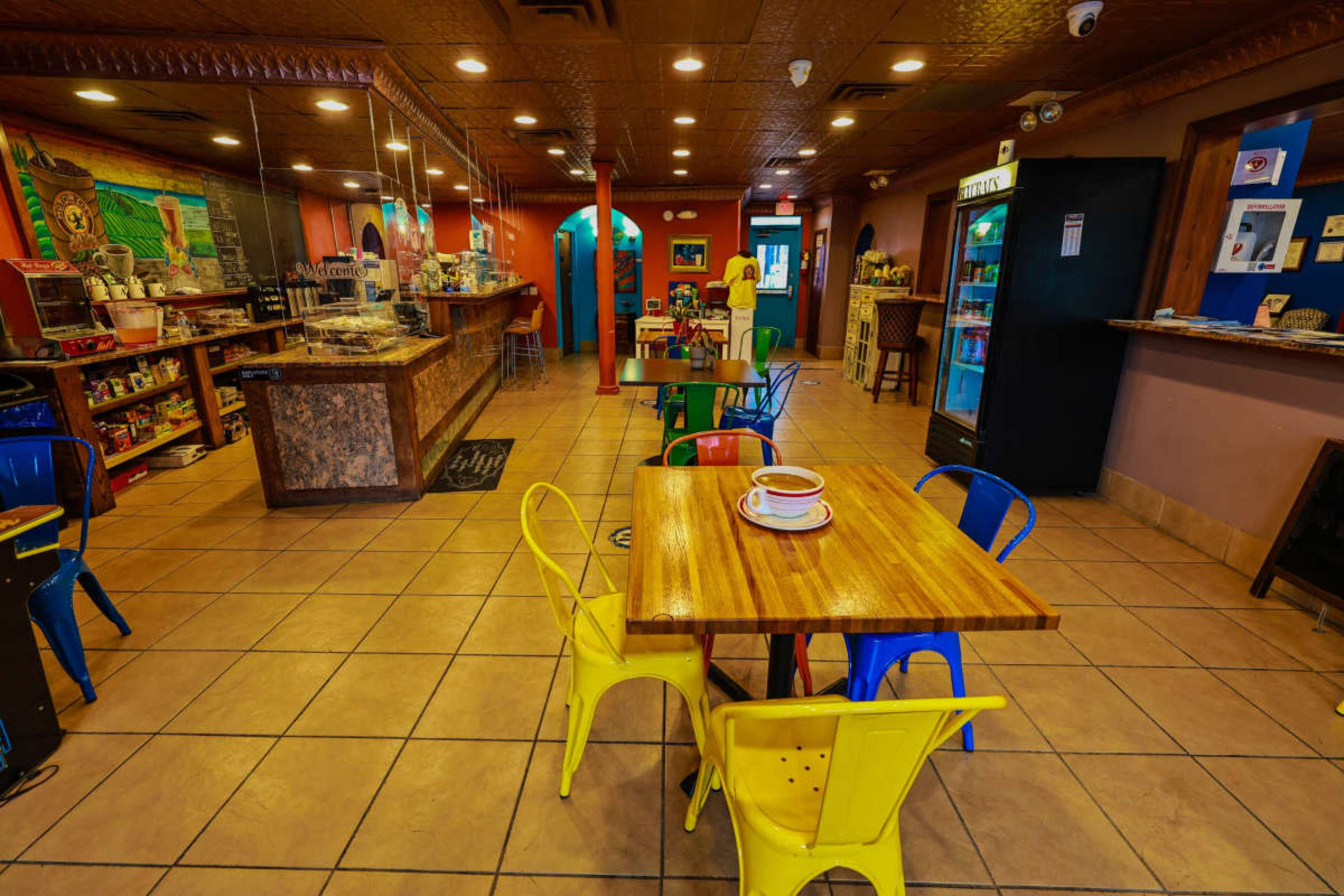 The image shows a brightly colored café interior with tables and chairs arranged on tiled floors, featuring a coffee counter in the background.