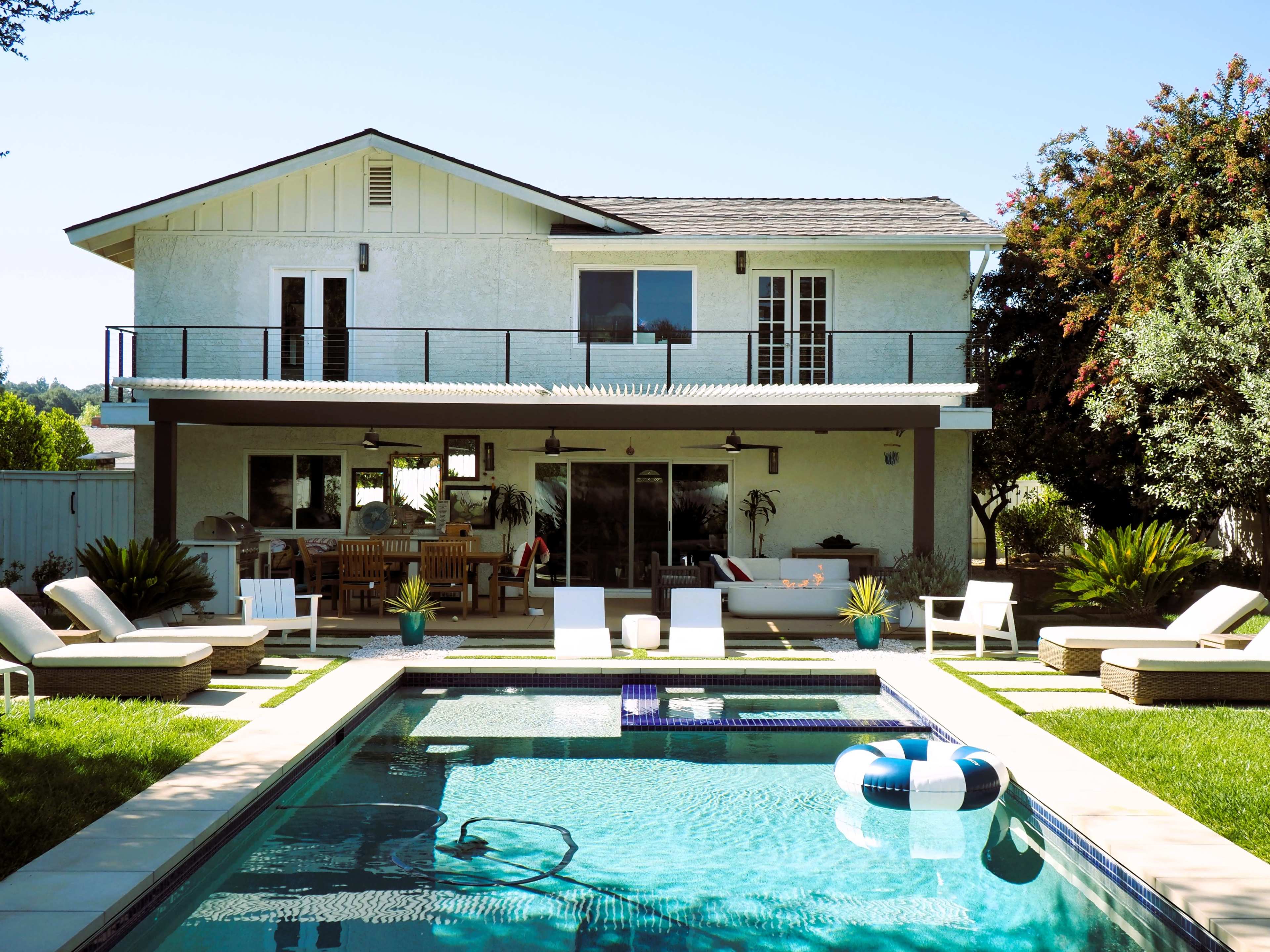A two-story house with a pool and patio furniture is surrounded by greenery in a residential backyard.