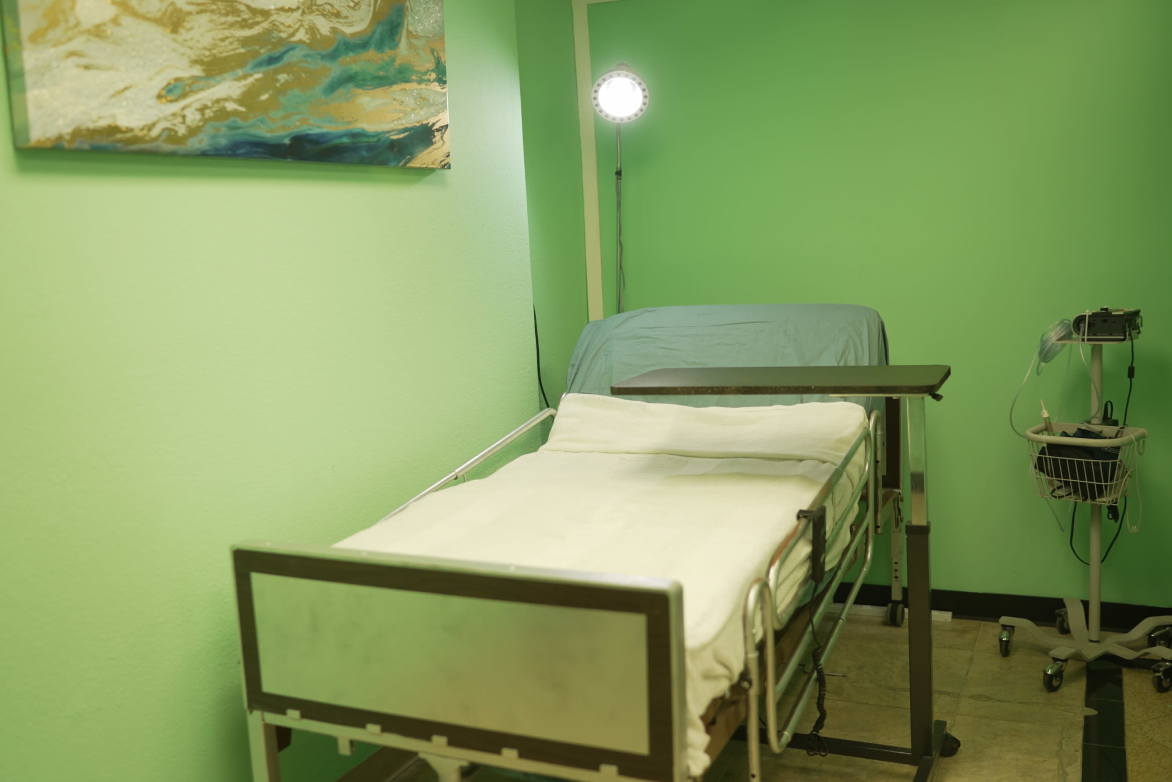 The image shows a hospital room with a vacant hospital bed covered in a white blanket, and a small medical cart beside it.