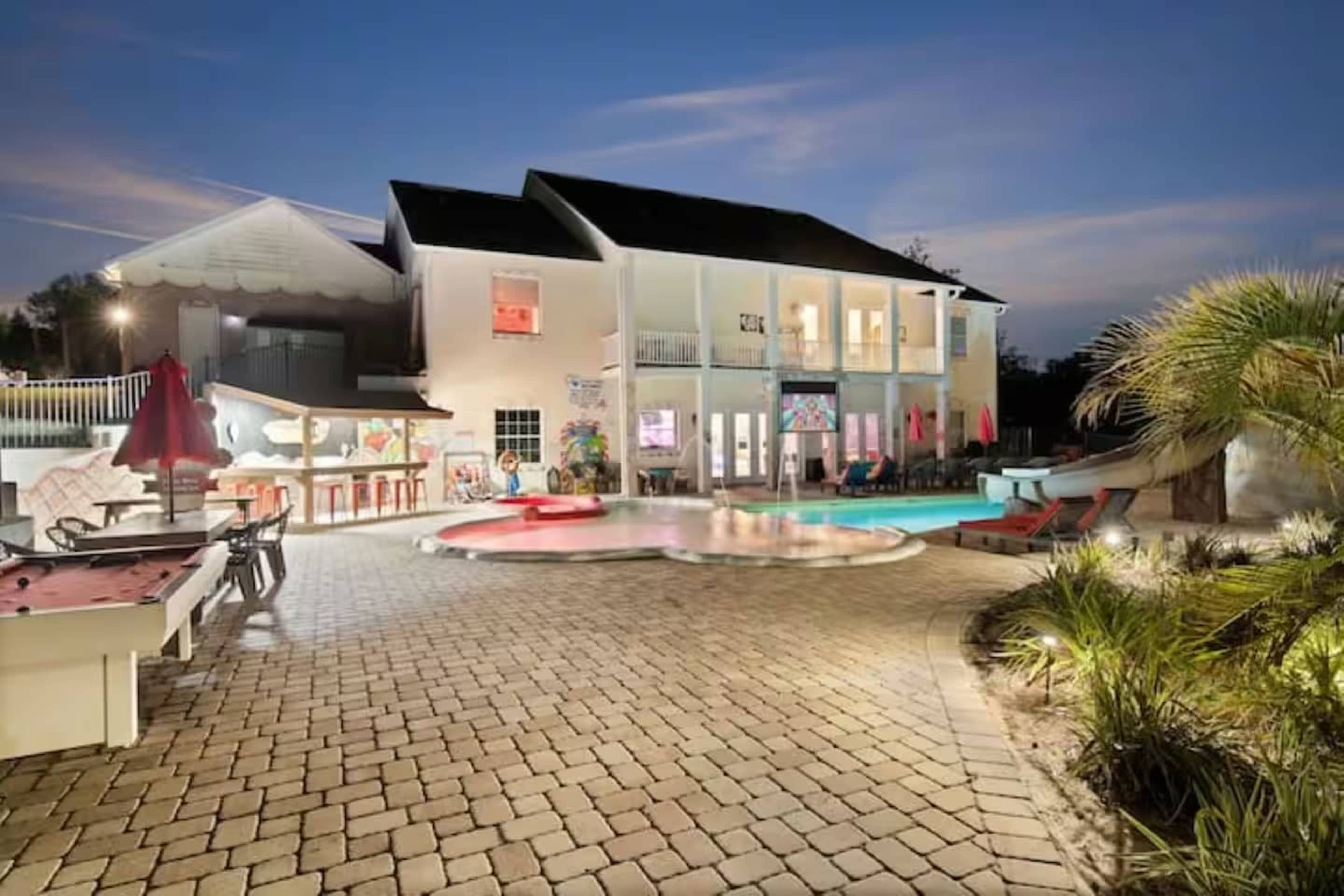 A large house with multiple balconies overlooks a pool surrounded by a paved patio and lounge areas, framed by palm trees and outdoor seating.