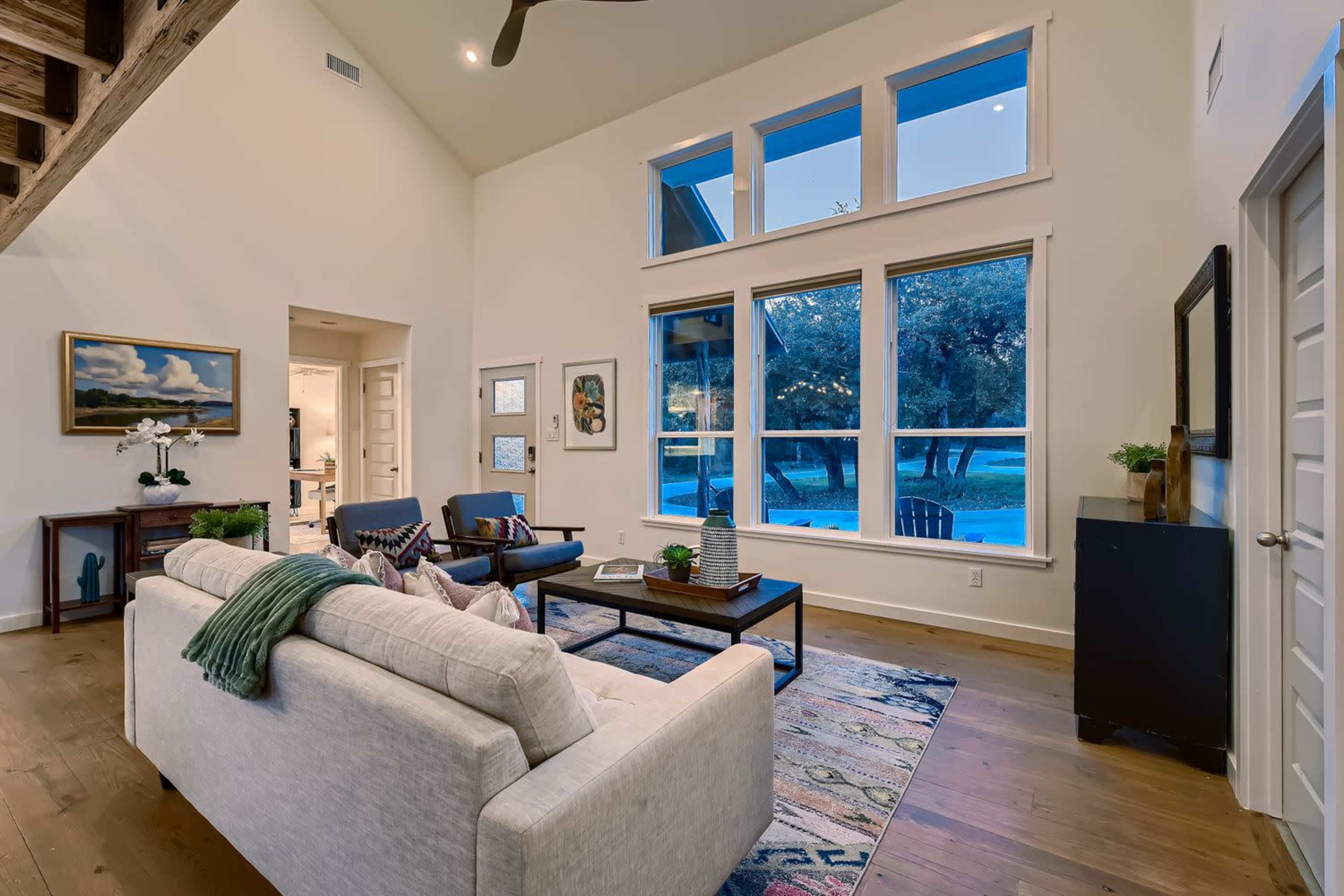 A spacious living room features a light-colored sofa, a coffee table, and large windows overlooking a wooded area.