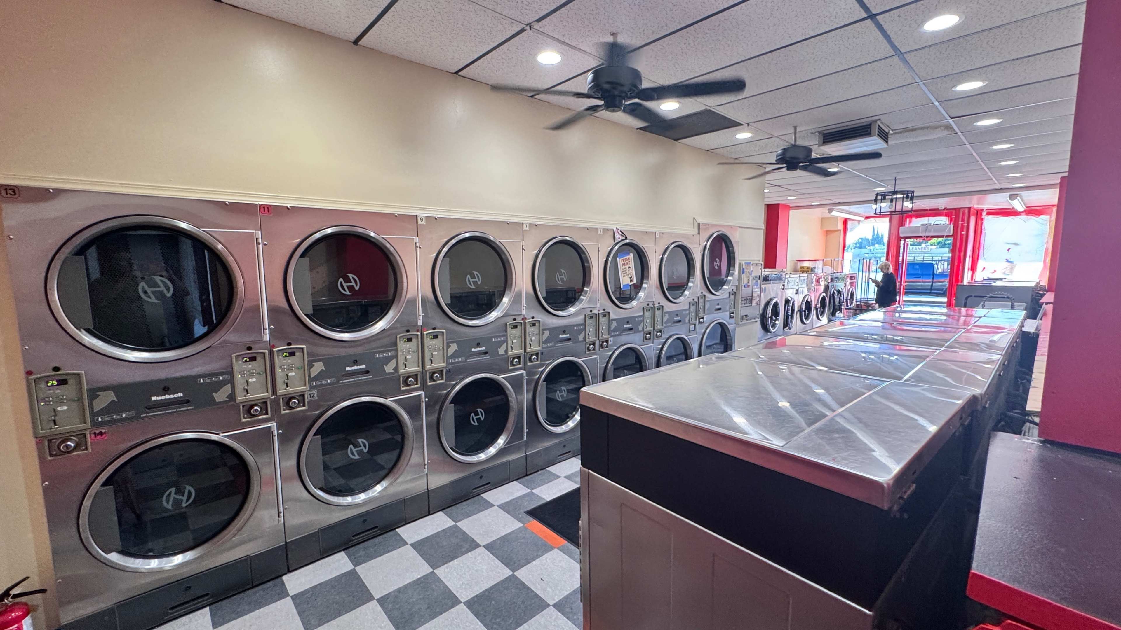 The image shows the interior of a laundromat with rows of washing machines and dryers along one wall, and a counter area on the opposite side.