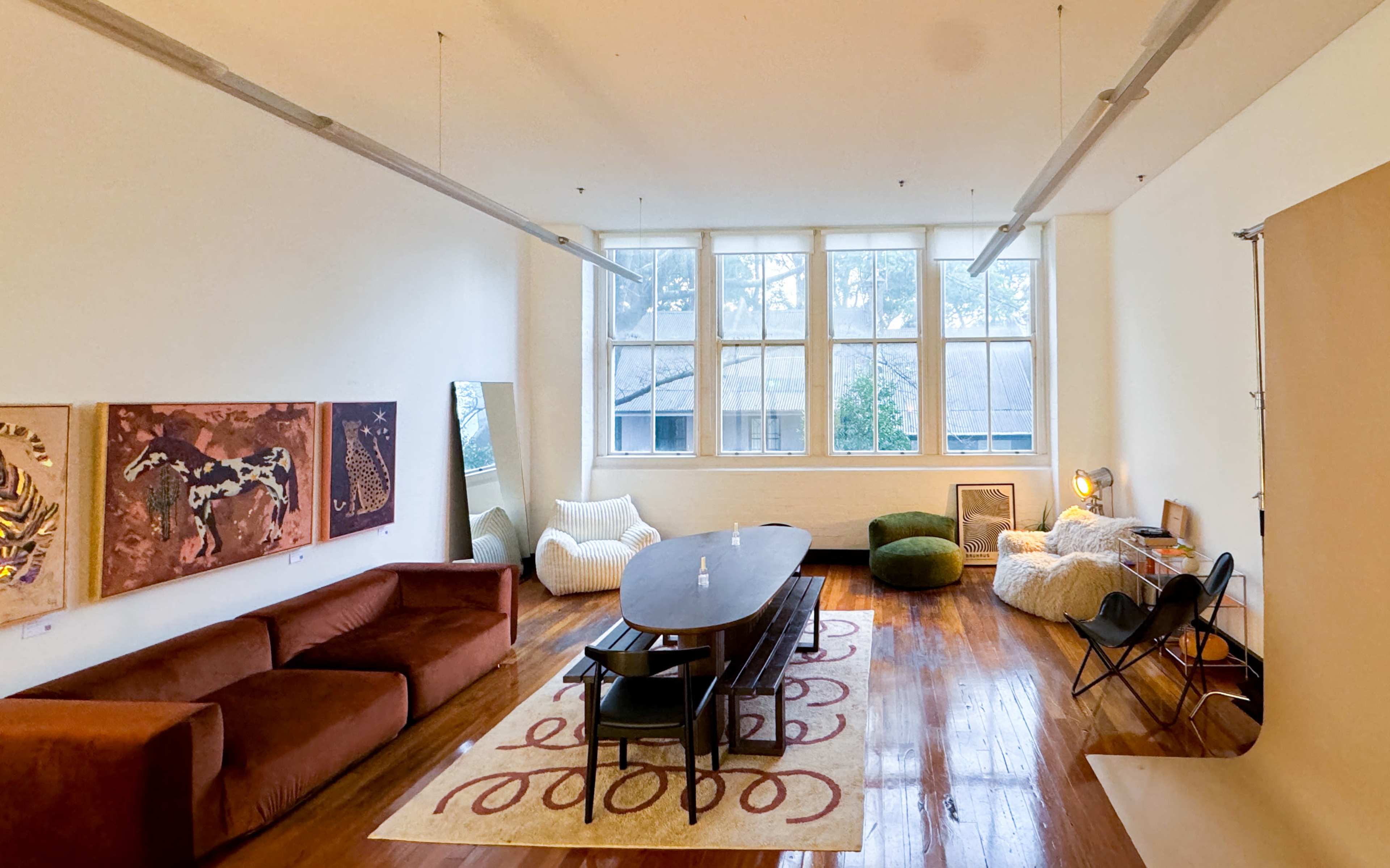 The image shows a spacious living area featuring a long table, a dark couch, bean bag chairs, large windows, and various artworks on the walls.