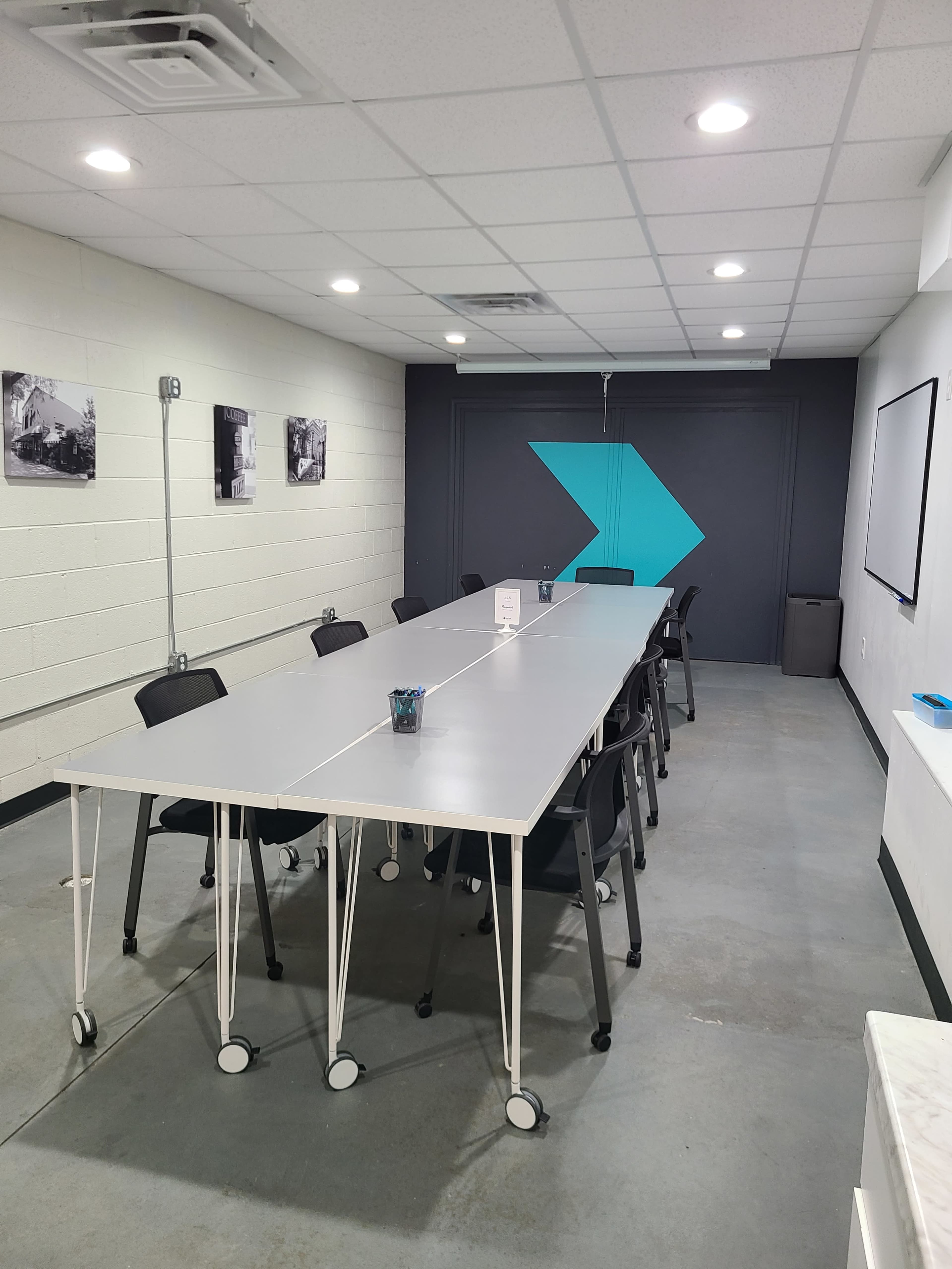 A long, modern conference table is set up in a well-lit room with a gray and teal accent wall, surrounded by black chairs.