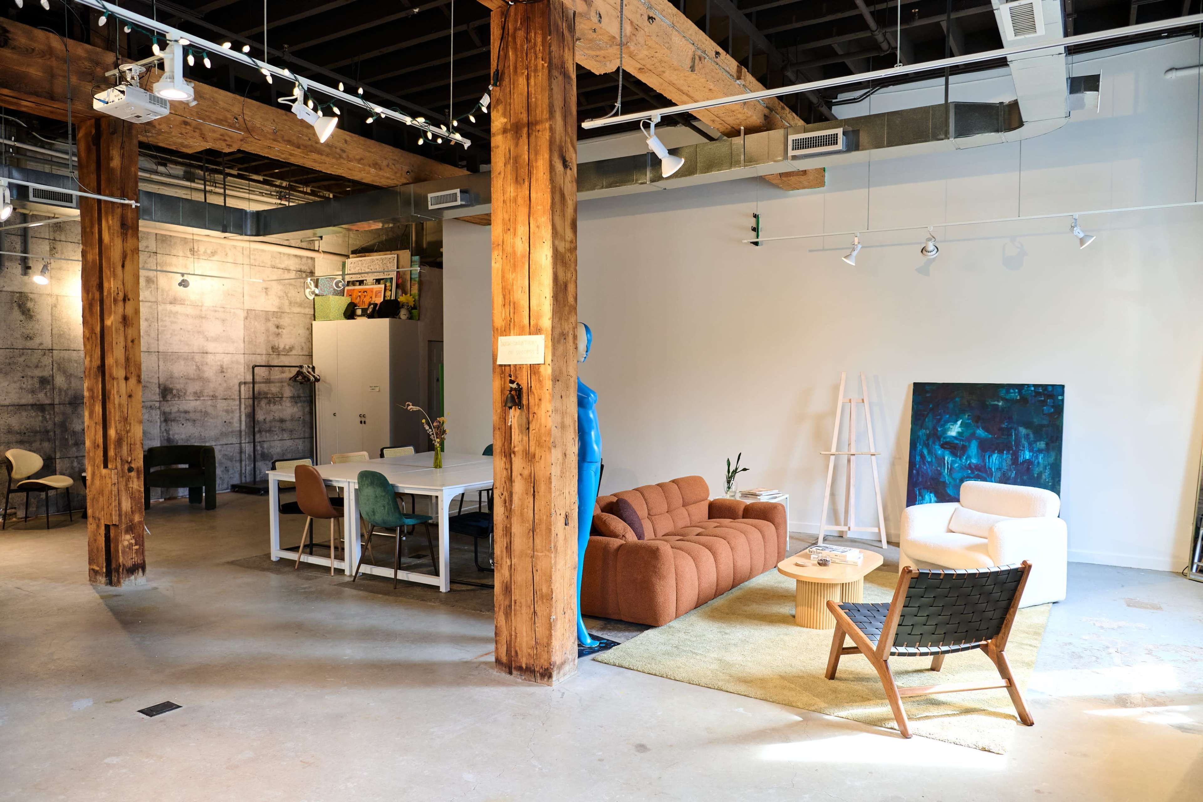 The image shows a spacious interior featuring a seating area with a brown sofa, a white chair, and a wooden coffee table, alongside a dining table and green chairs under exposed wooden beams.