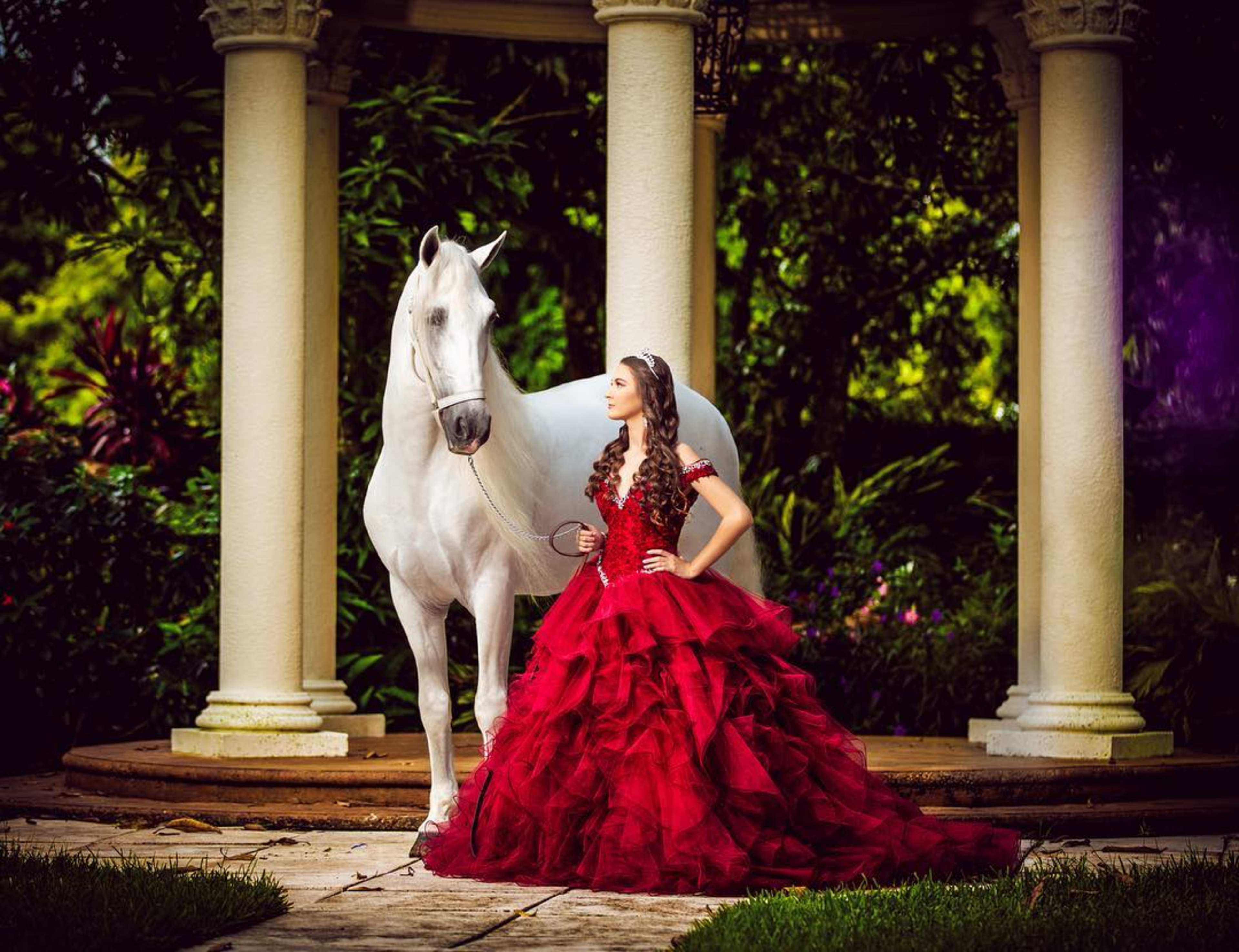 A young woman in a flowing red gown stands beside a white horse near a columned structure in a lush garden.