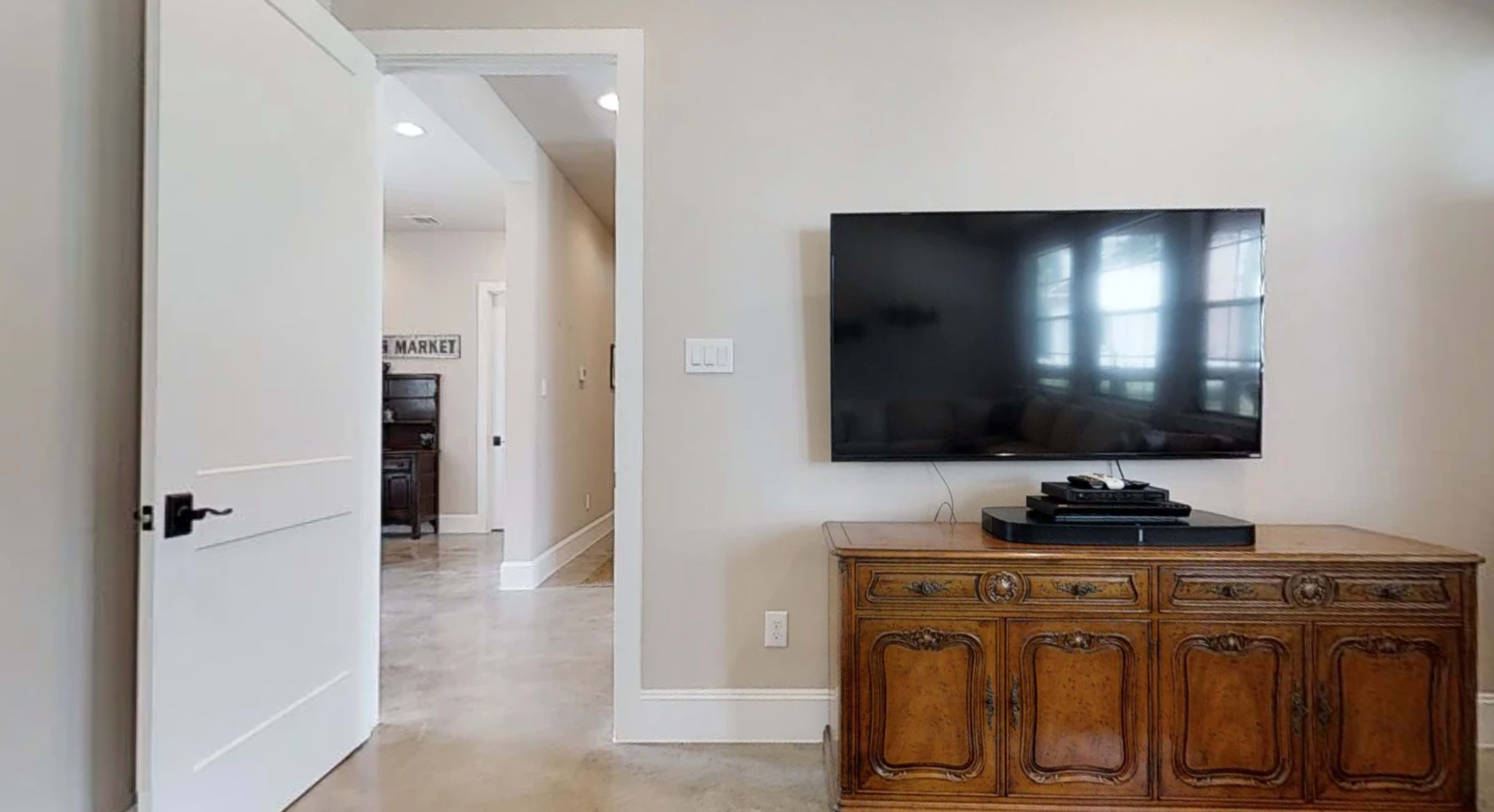 A flat-screen TV is mounted on a wall above a wooden media console in a well-lit room, with a white door leading to another space in the background.