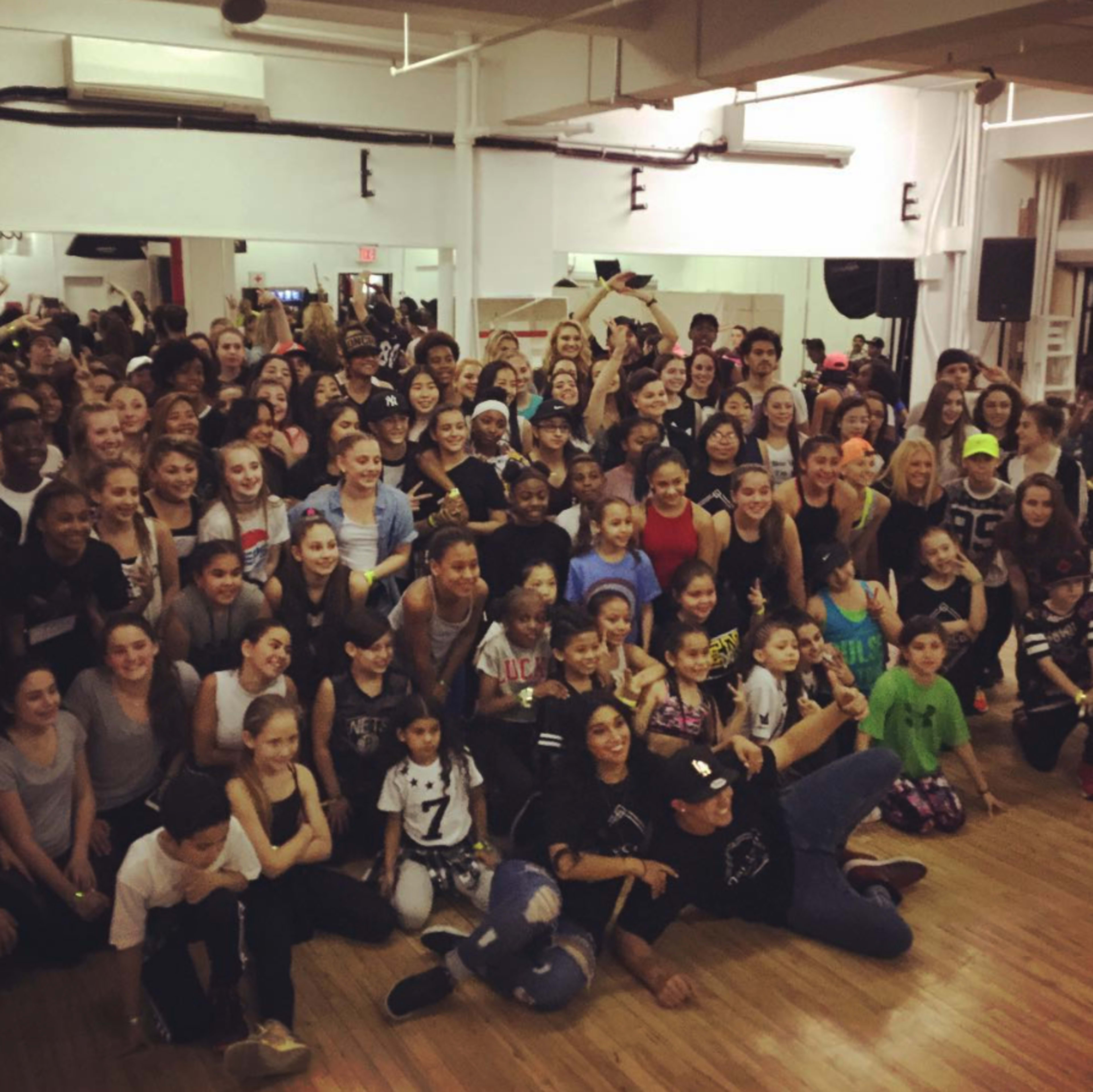 A large group of people, including children and adults, gathers for a dance class in a studio with wooden floors and mirrors.