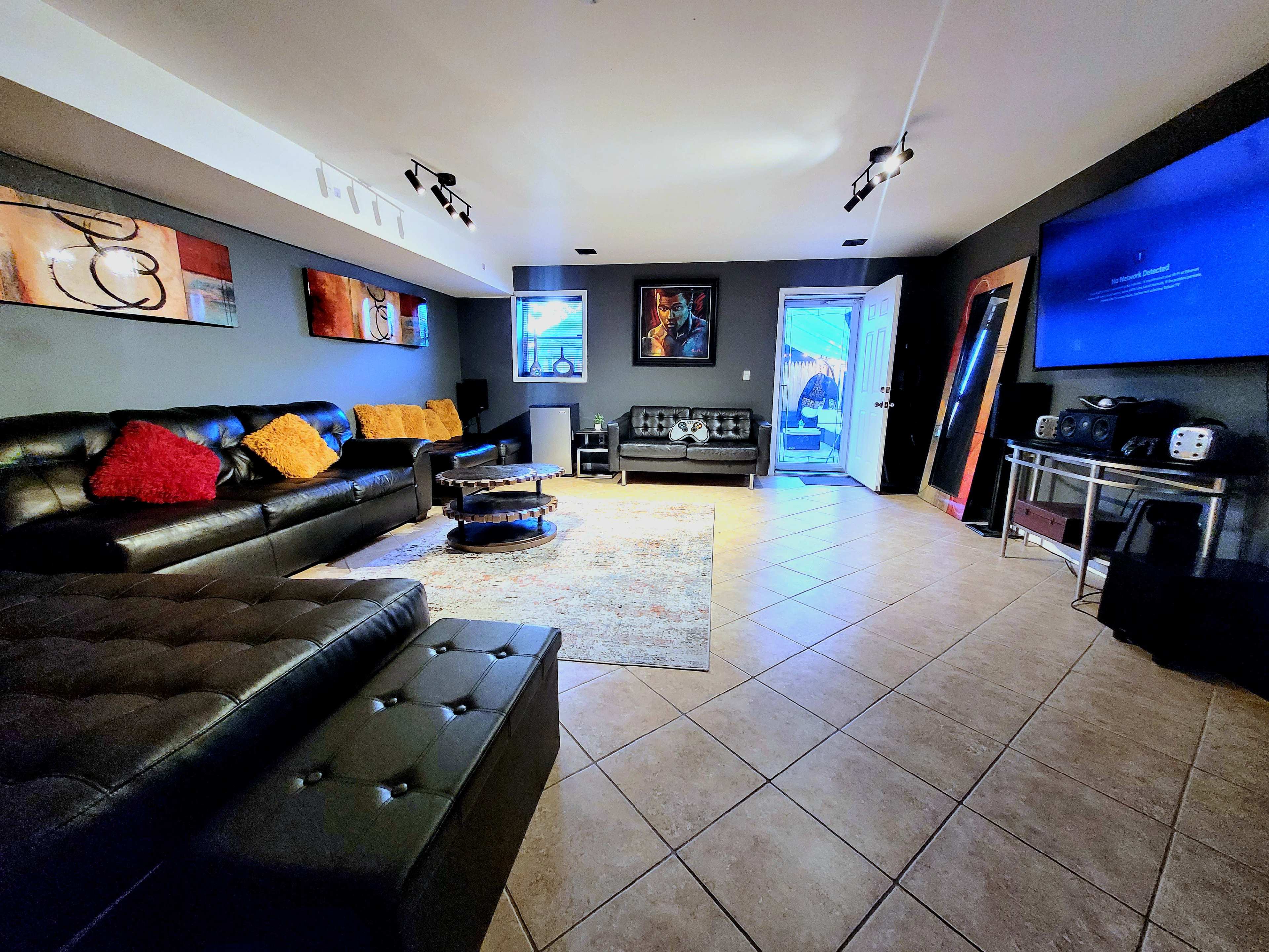 A modern living room features a leather sectional sofa, a rug on tiled flooring, and a large television mounted on the wall, with a door leading outside.