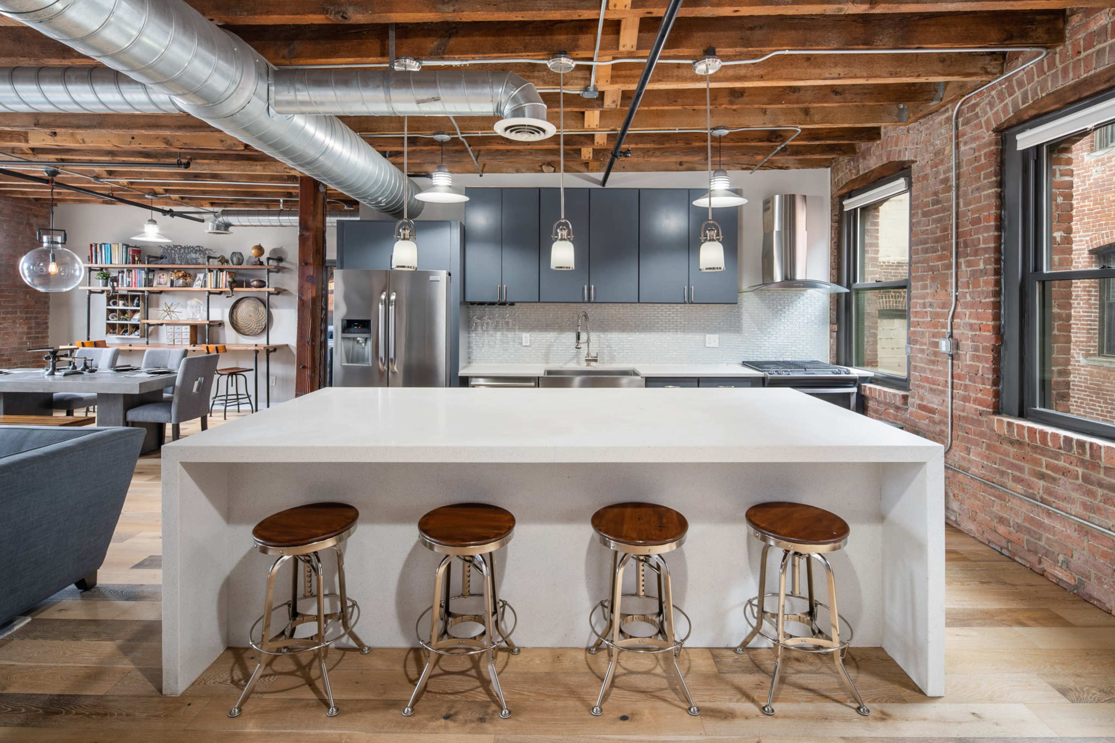 The image shows a modern kitchen area with a large white countertop island and four metal stools, adjacent to exposed brick walls and wooden ceiling beams.