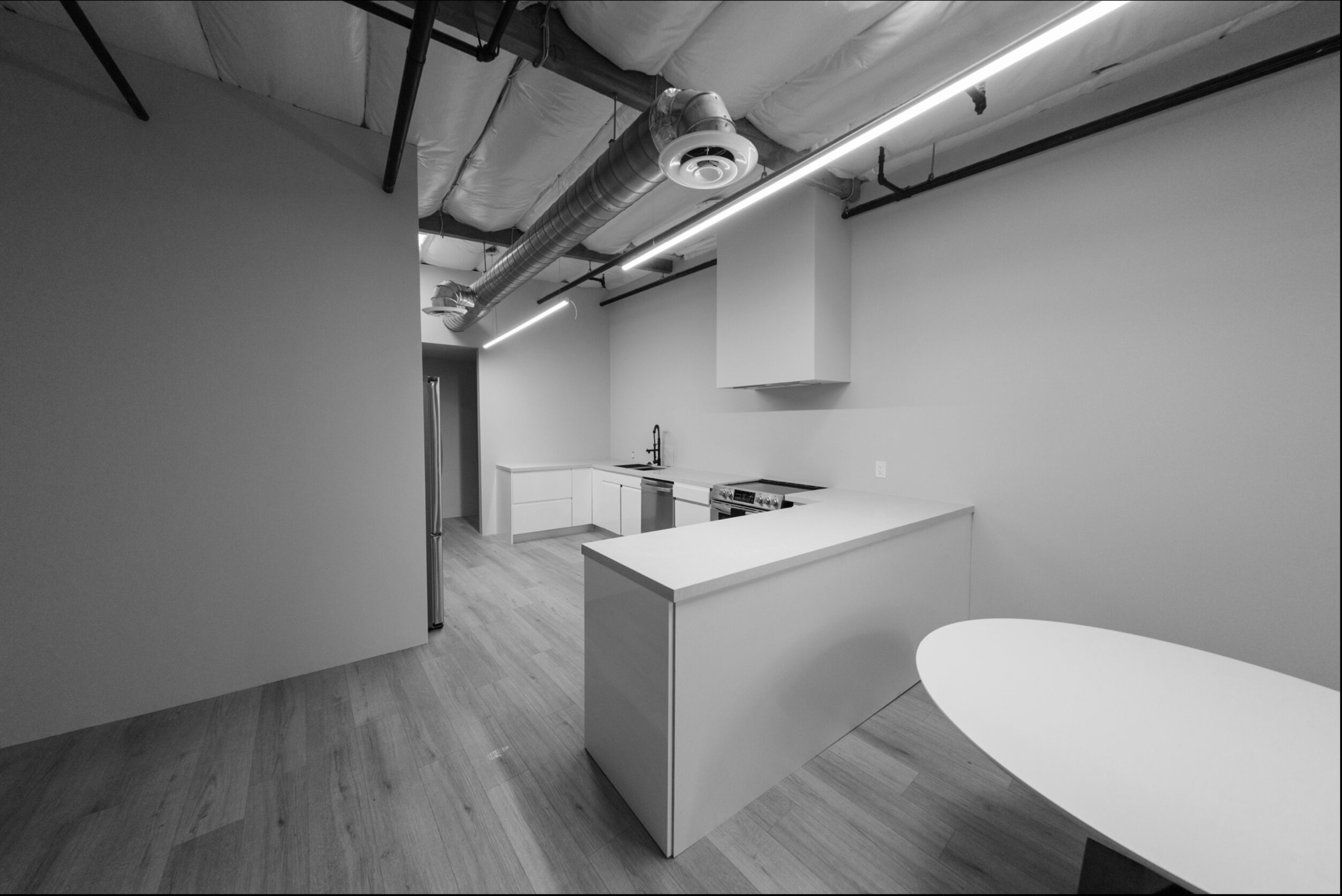 The image shows a spacious, minimalistic kitchen area featuring a central island, white cabinetry, and modern appliances, with a light wood floor and exposed ceiling ducts.