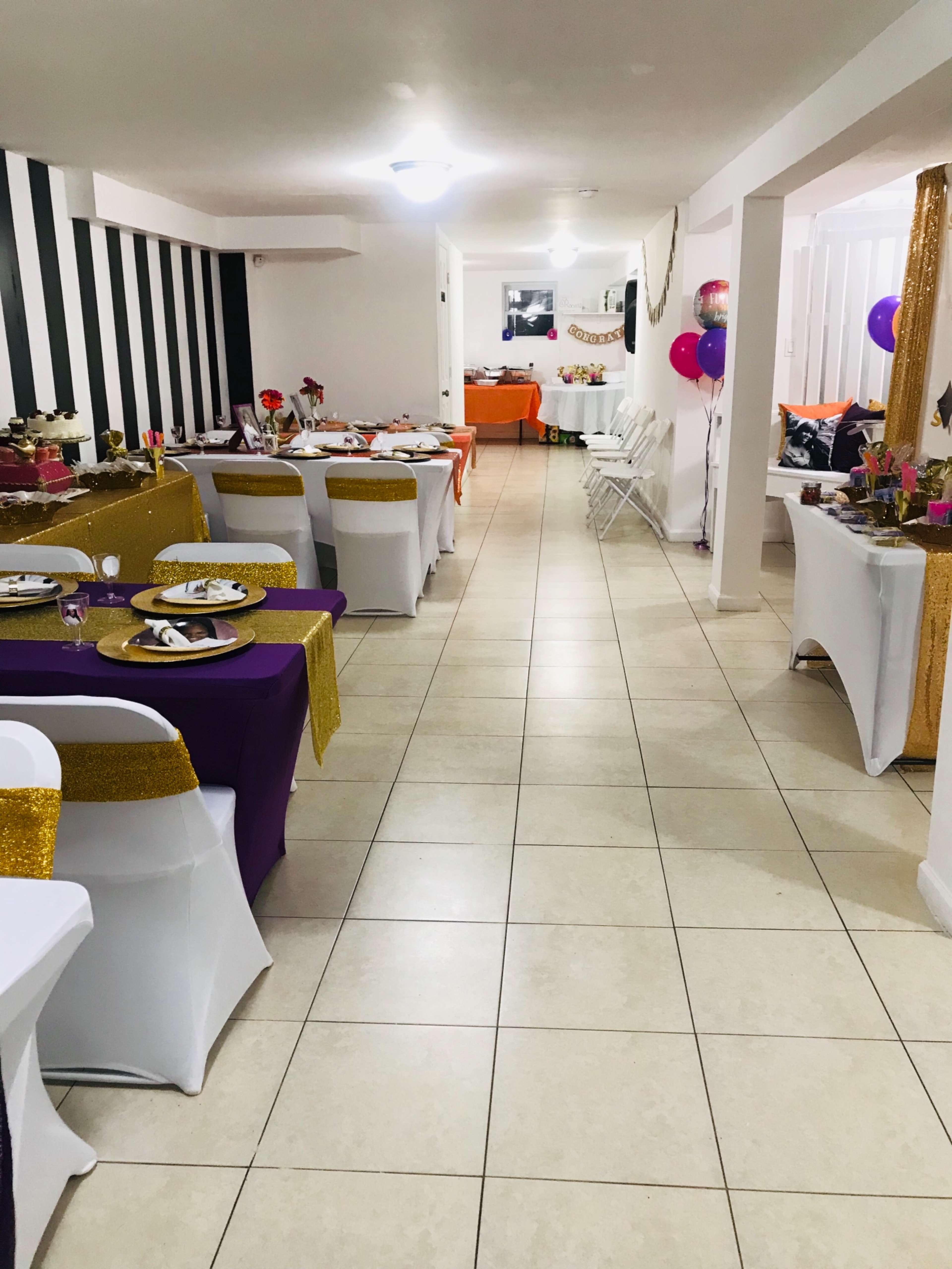 A brightly decorated event space with tables covered in various colored cloths set up along the walls, flanked by chairs and balloons.
