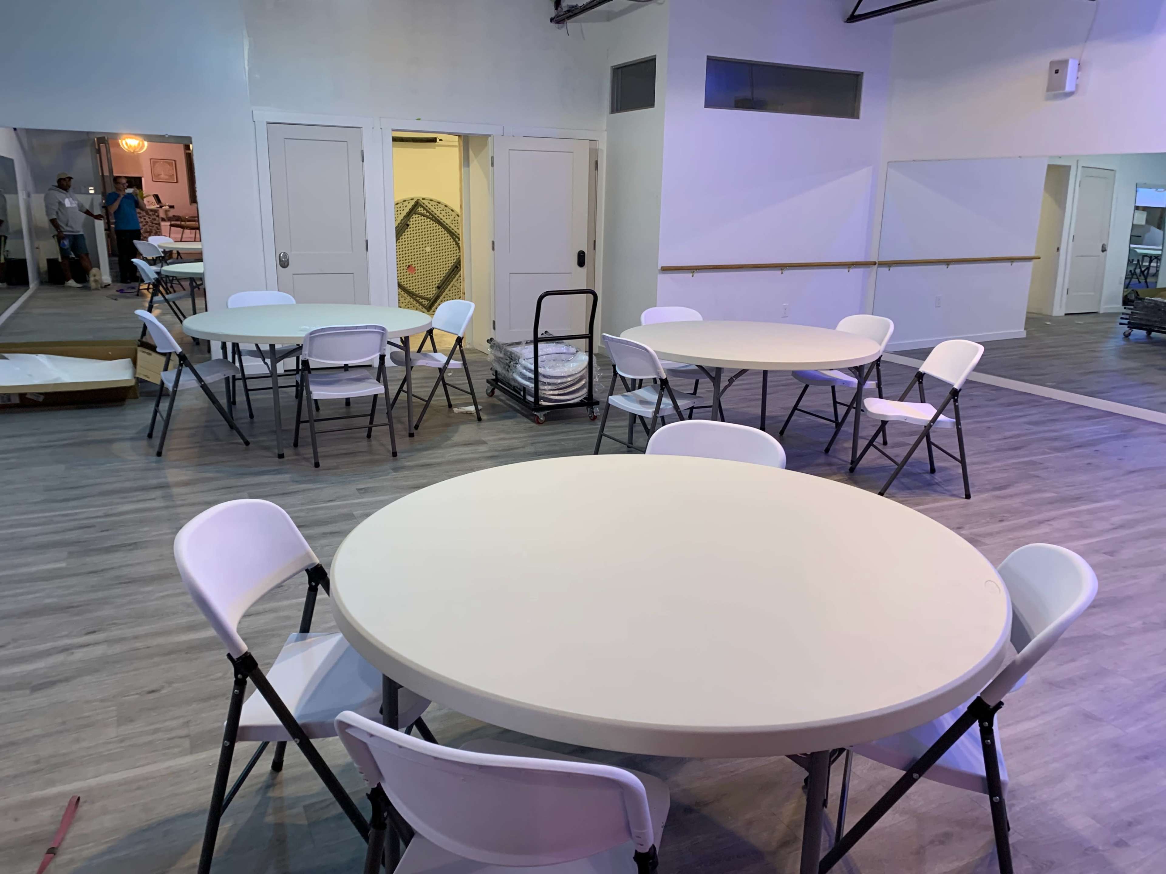 A spacious room with multiple round tables and folding chairs arranged for an event, with mirrors and a doorway visible in the background.