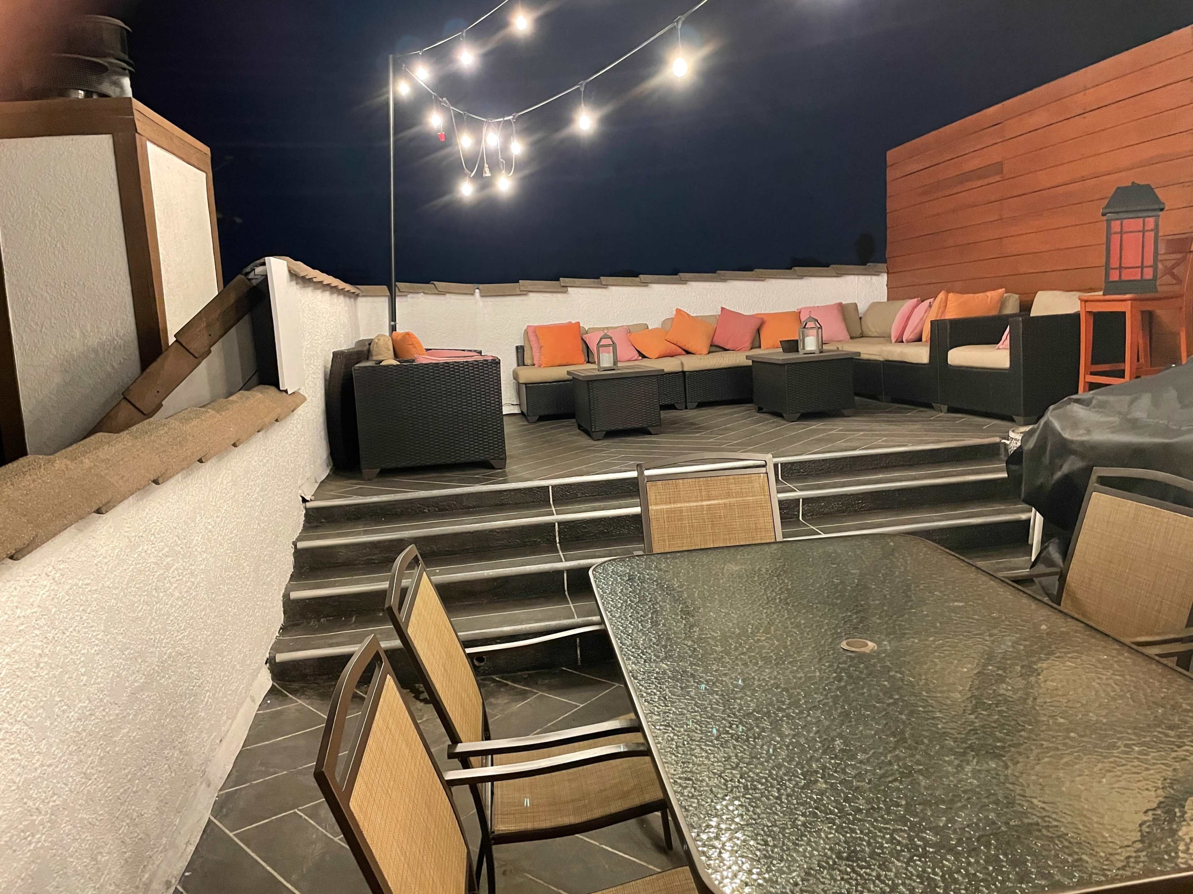 A rooftop patio features a seating area with dark furniture and colorful cushions, illuminated by string lights above.
