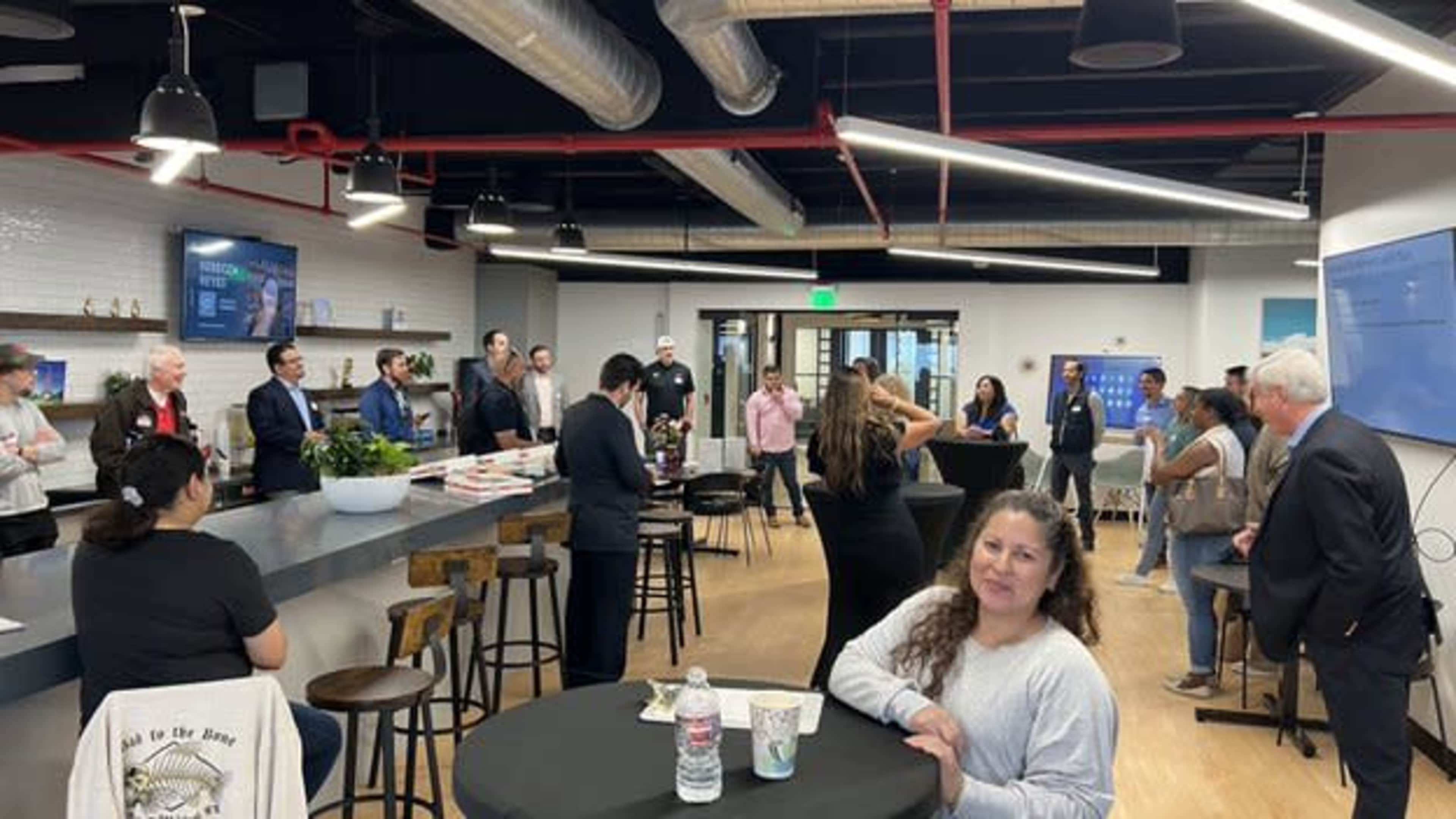 A group of people are gathered in a modern office space with high tables and a bar area, engaging in conversation during an event.