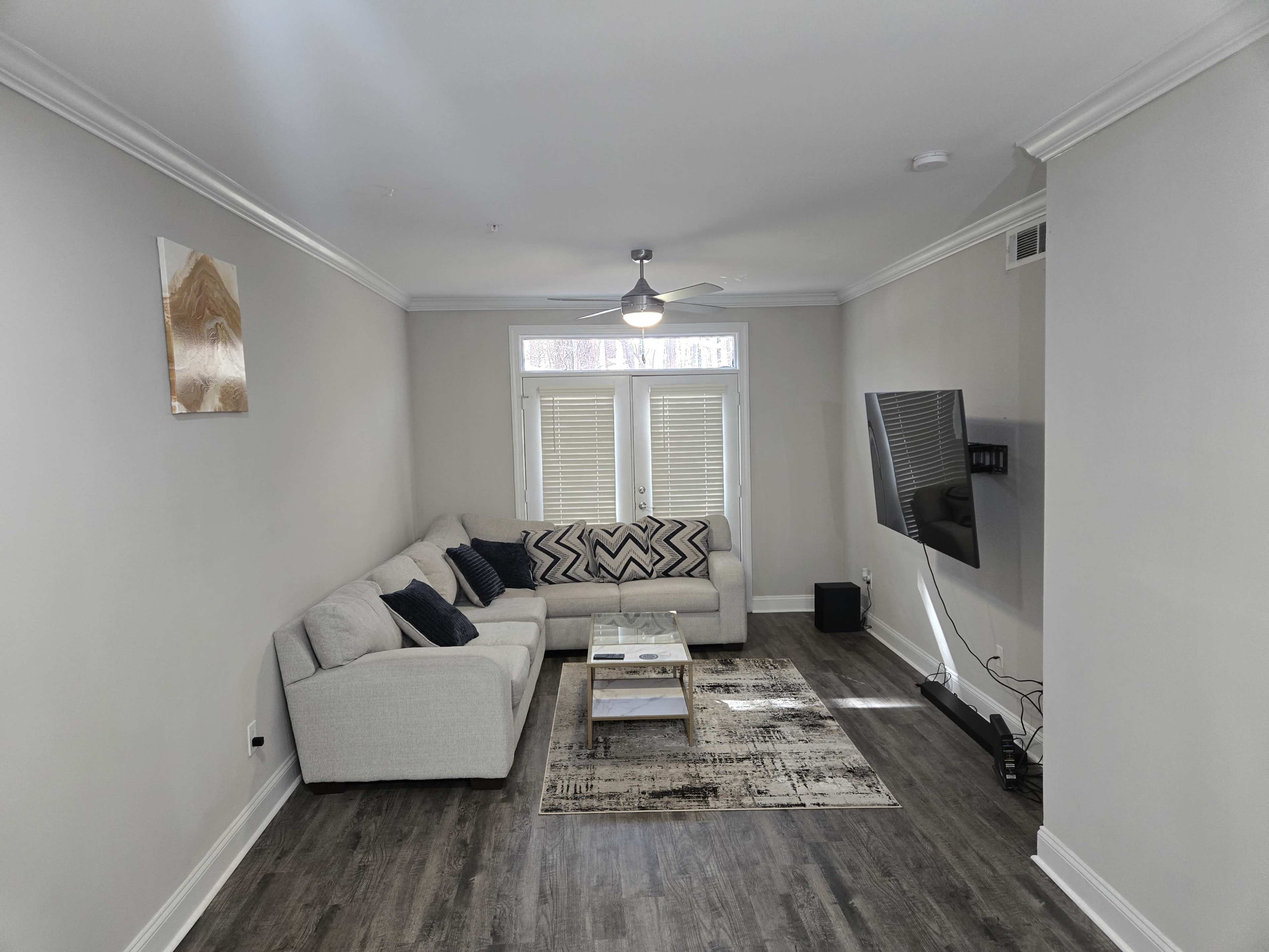 The image shows a modern living room with a gray sectional sofa, a coffee table, a flat-screen TV mounted on the wall, and large windows with closed blinds.