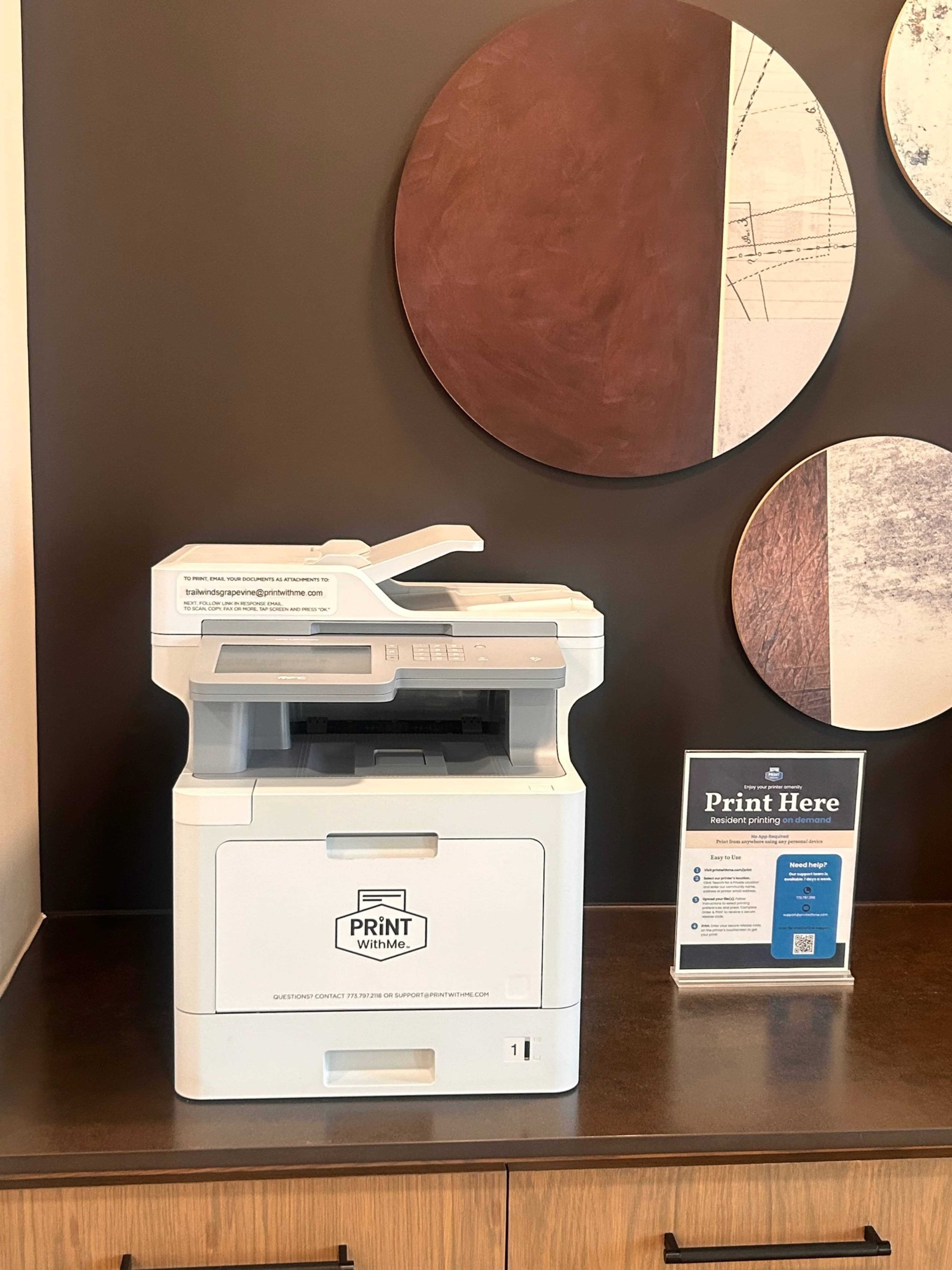 A printer is positioned on a wooden table with a sign labeled "Print Here" nearby, in front of circular wall decor.
