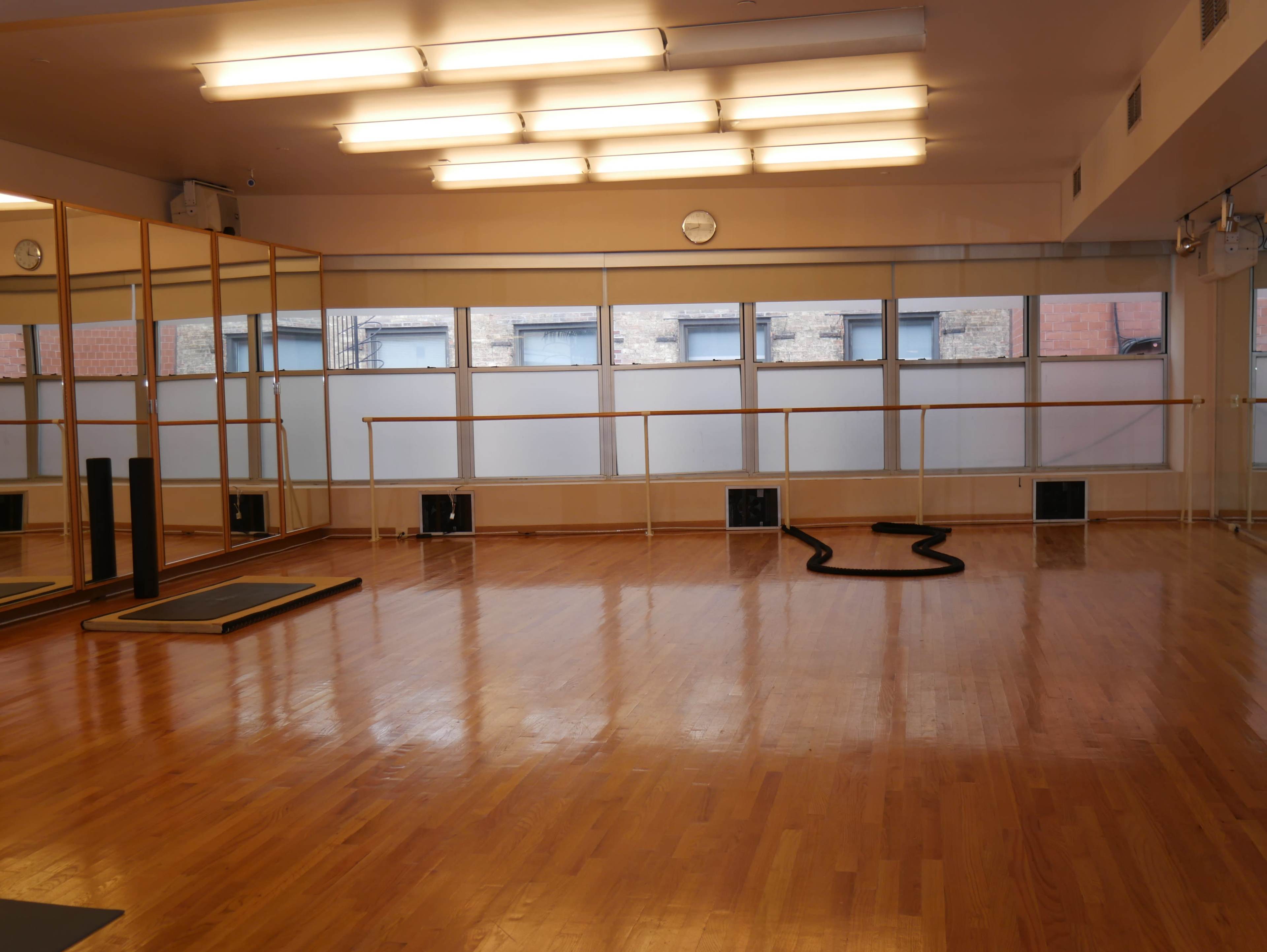 A spacious, empty dance or exercise studio with wooden flooring, large mirrors, and natural light coming through the windows.