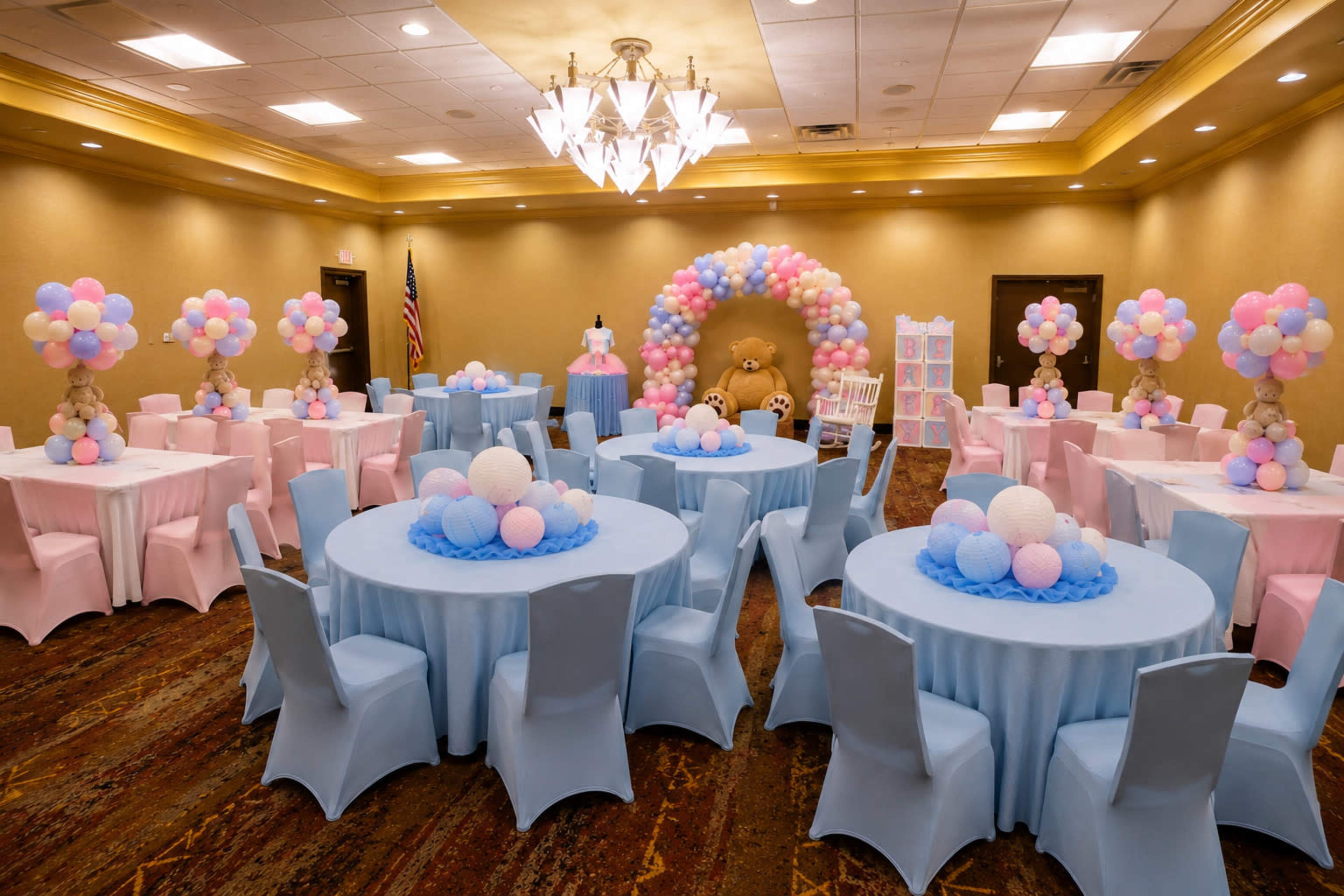 A banquet hall is decorated for a celebration, featuring round tables covered in light blue and pink fabrics, with balloon arrangements and a colorful backdrop.