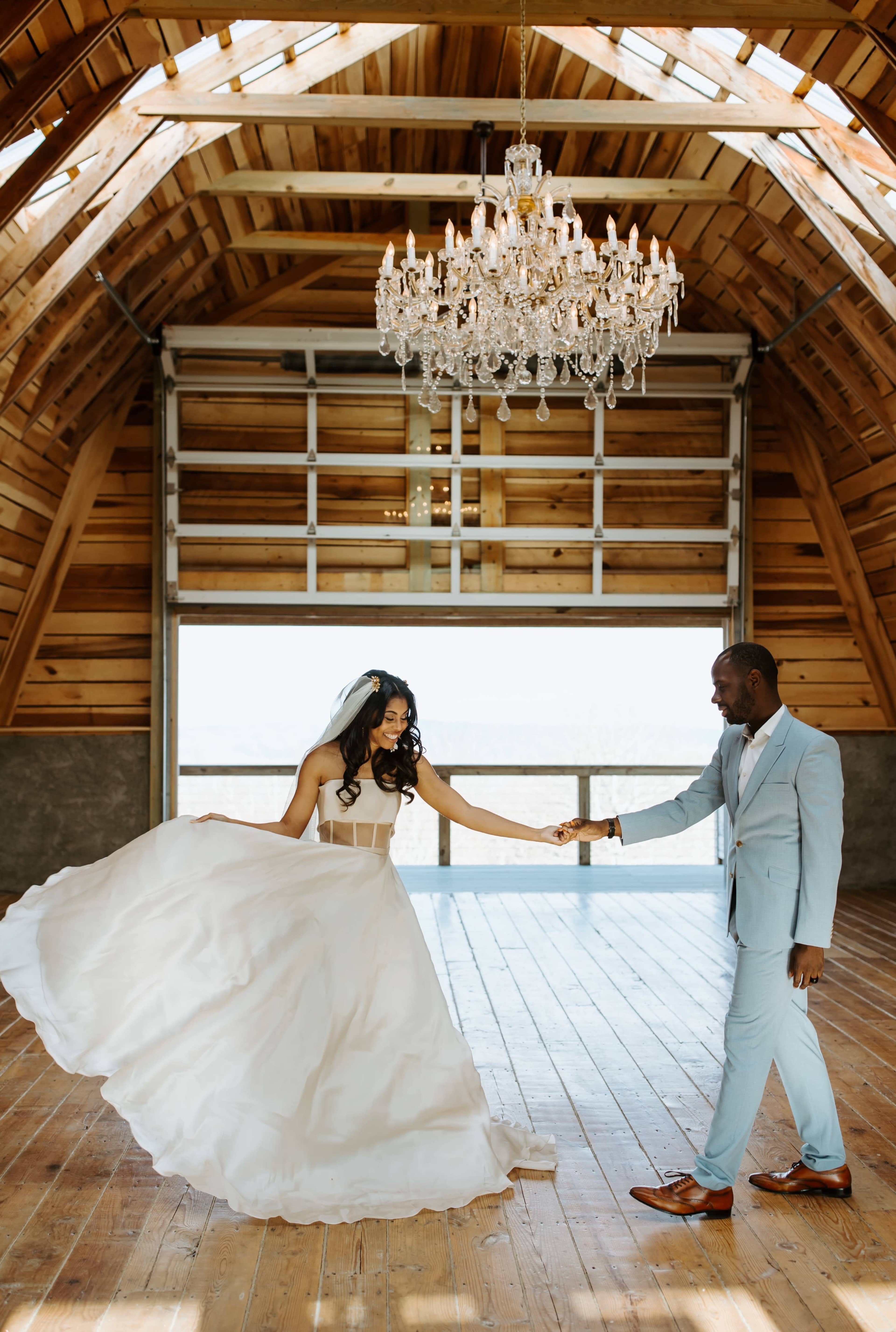 A couple holds hands in an elegantly designed barn venue with wooden beams and a chandelier overhead.