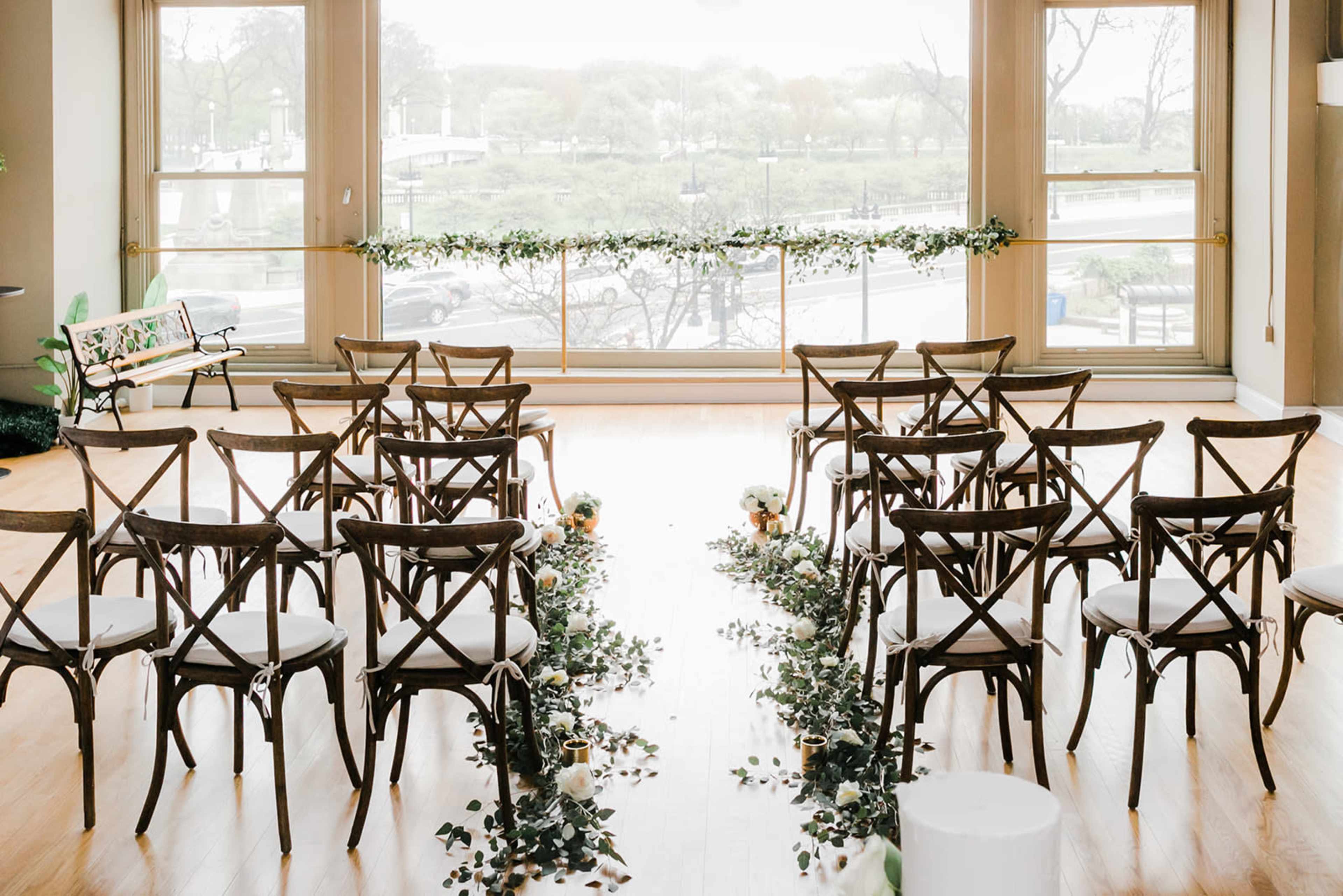 A wedding ceremony setup with rows of wooden chairs arranged in front of a decorative aisle made of greenery, all facing a large window with a view of an outdoor setting.