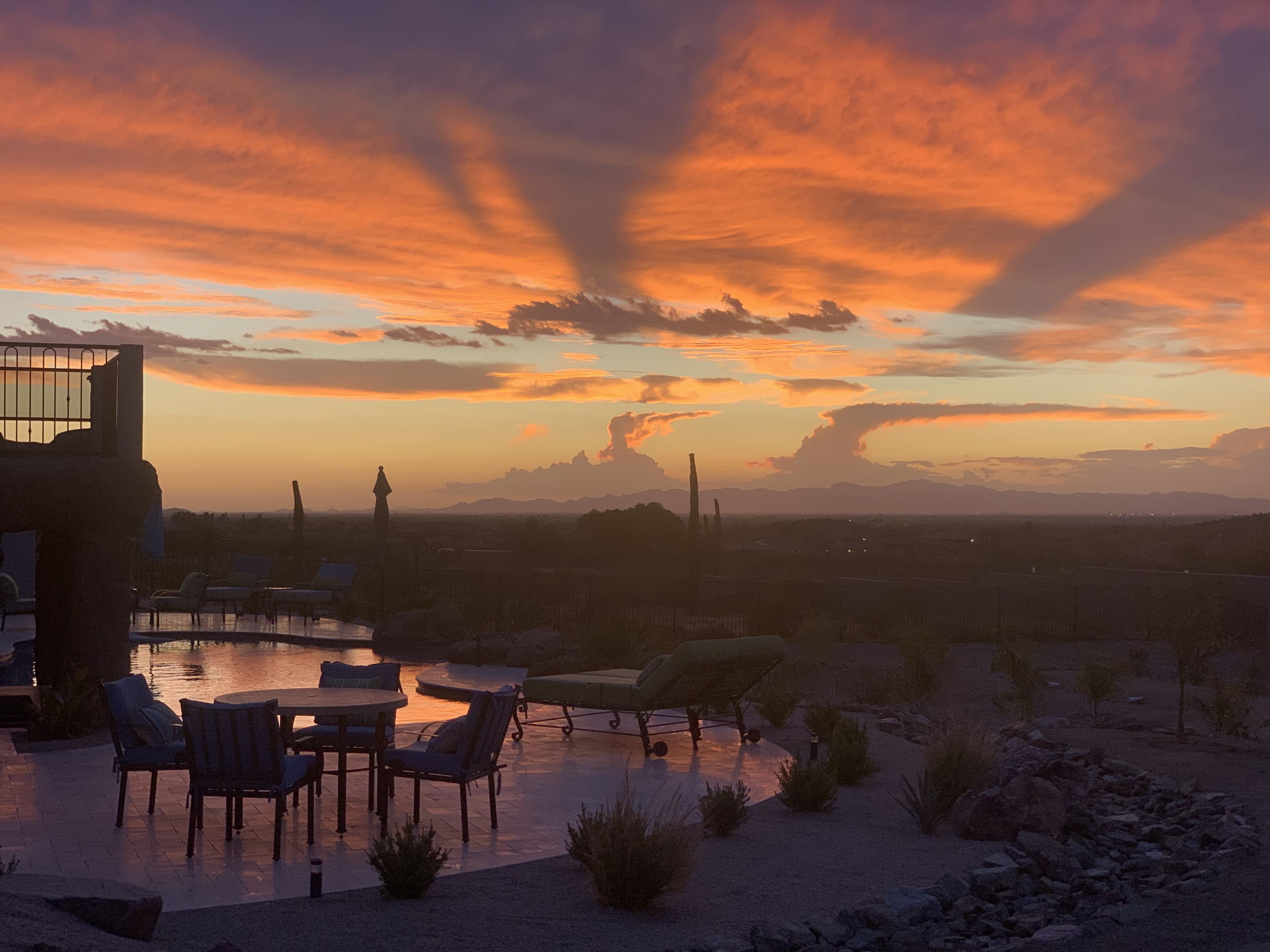 A sunset casts vibrant colors across the sky, reflecting off a pool surrounded by seating and desert vegetation.