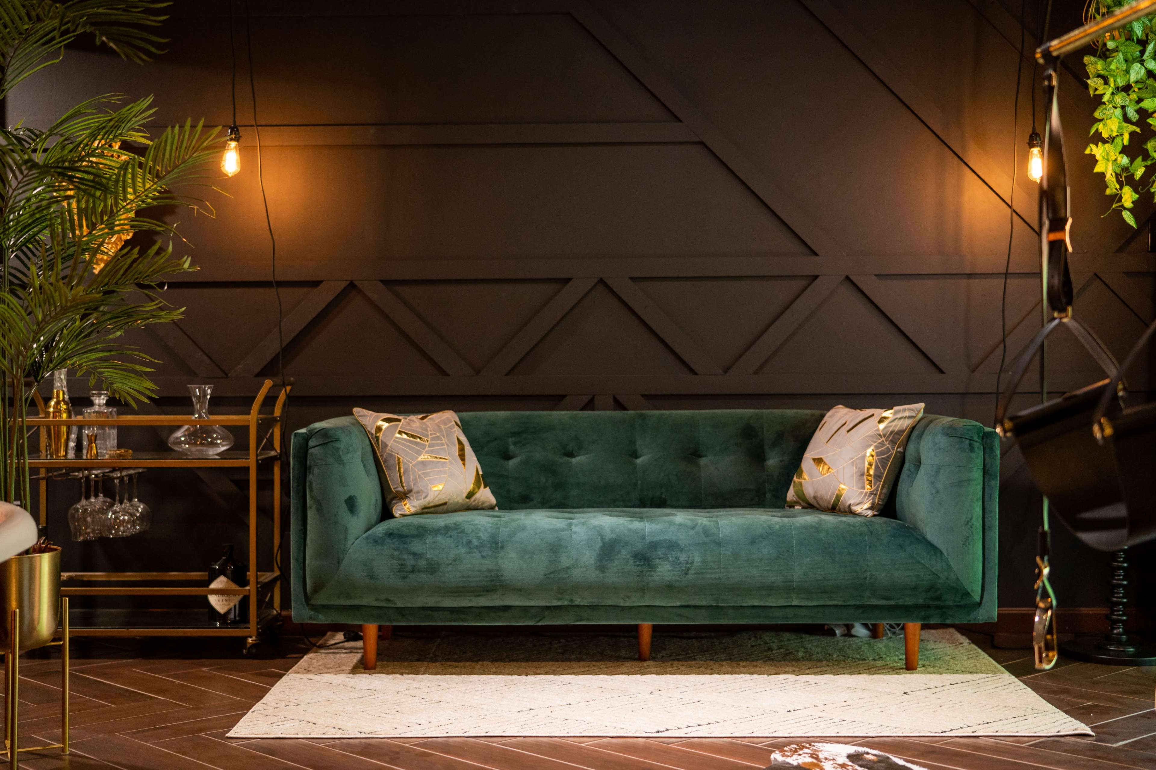 A green velvet sofa with decorative pillows placed against a dark wall, surrounded by indoor plants and vintage-style lighting.