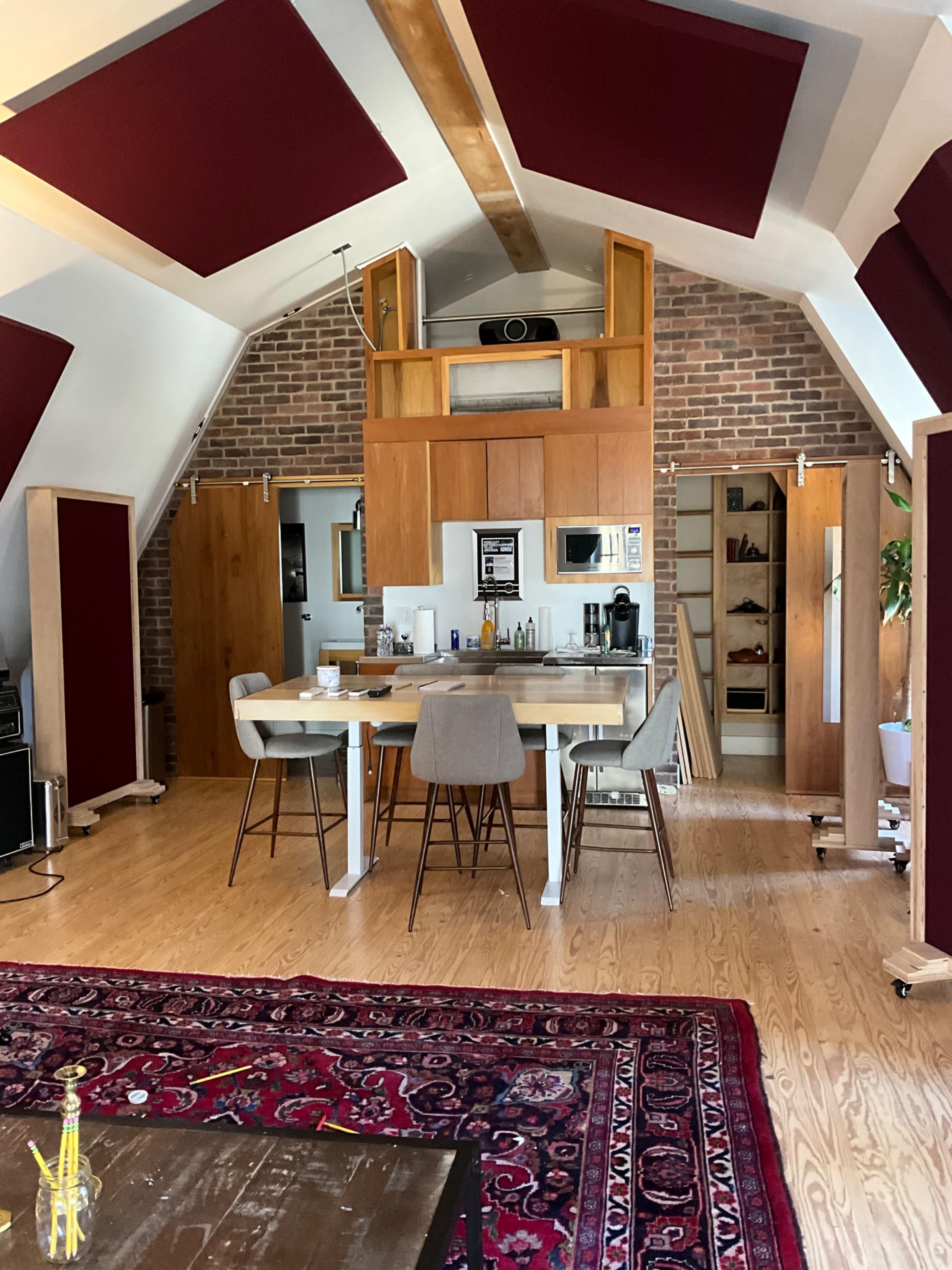 A spacious room with high ceilings featuring wooden beams, a dining table surrounded by chairs, and a kitchen area with wooden cabinets and a brick wall.