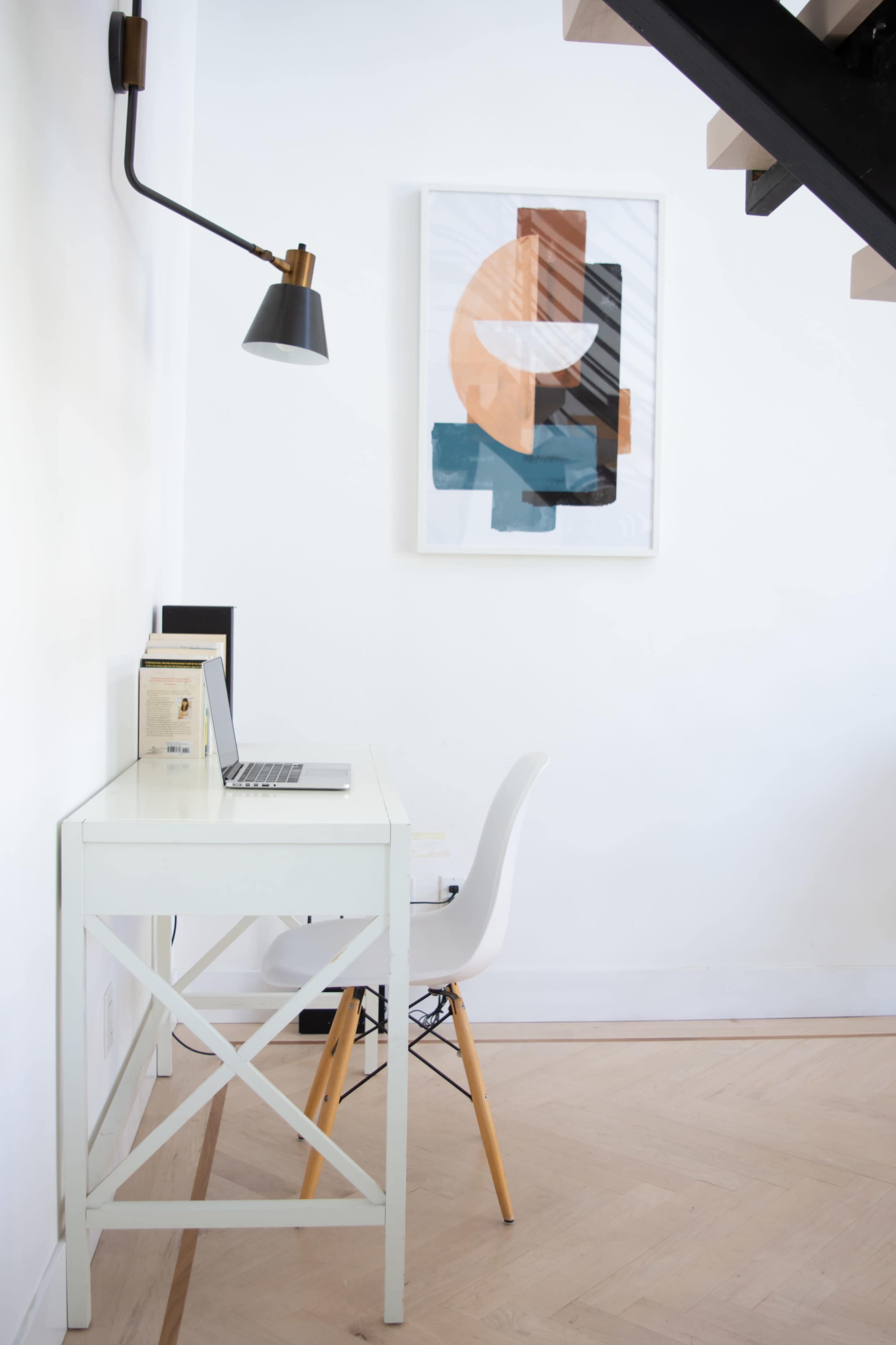A minimalist workspace features a white desk with a laptop, a chair, and a geometric art piece on the wall, situated beneath a staircase.