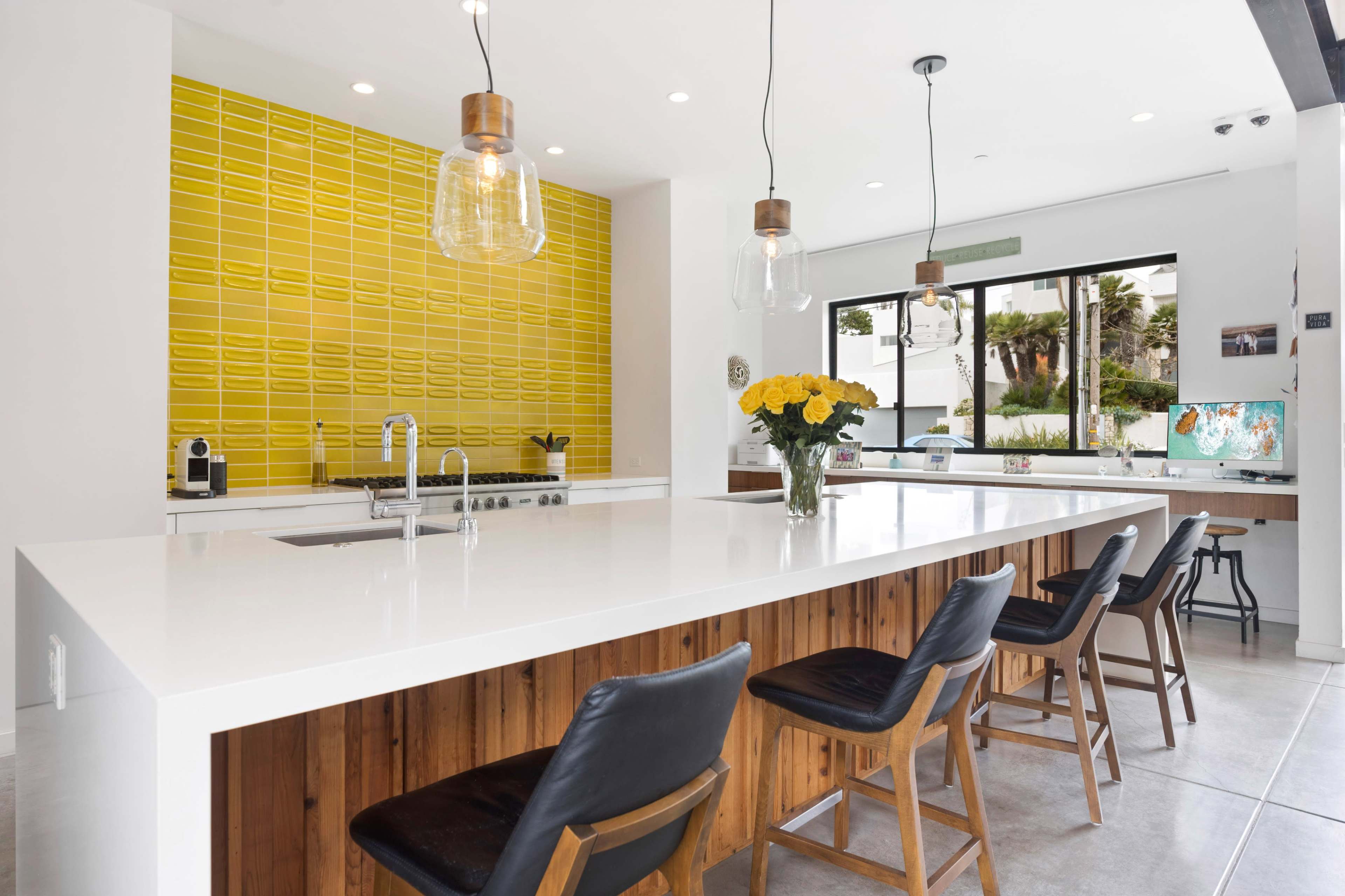 A modern kitchen with a large white island counter, wooden bar stools, and a bright yellow tiled backsplash.