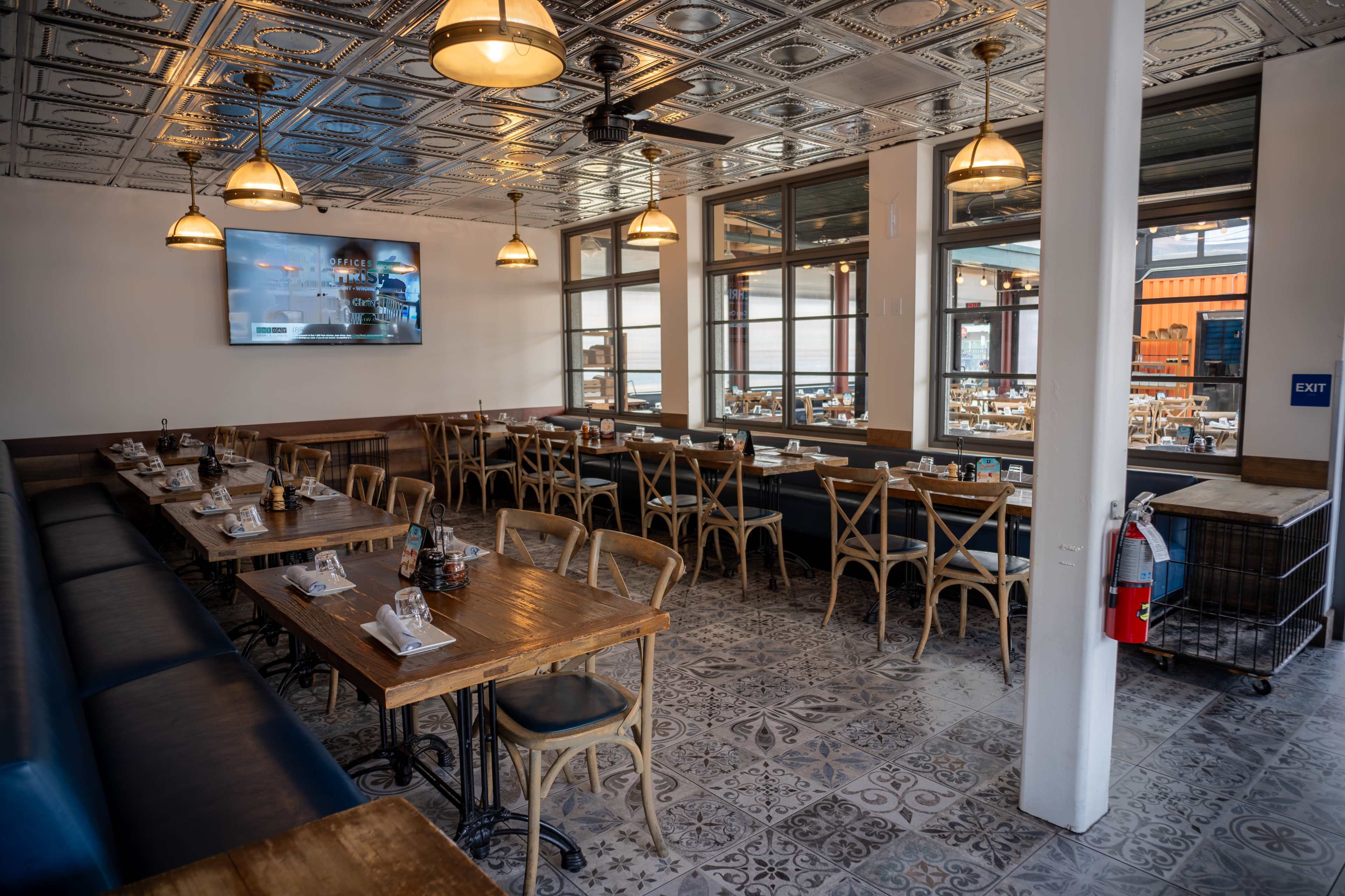The image shows an interior of a restaurant featuring wooden tables, chairs, and a patterned tiled floor, with a television mounted on one wall.