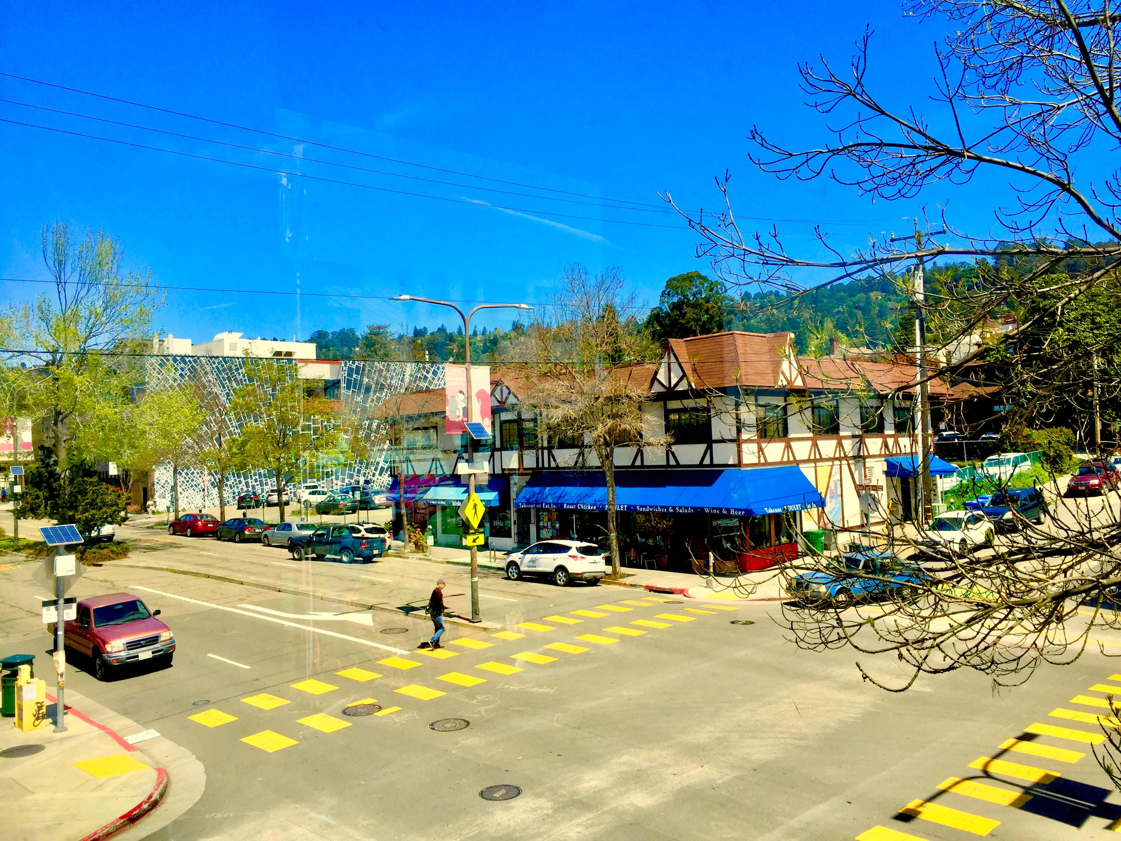 The scene shows a busy intersection with a person walking across a crosswalk, surrounded by various parked cars and buildings under a clear blue sky.