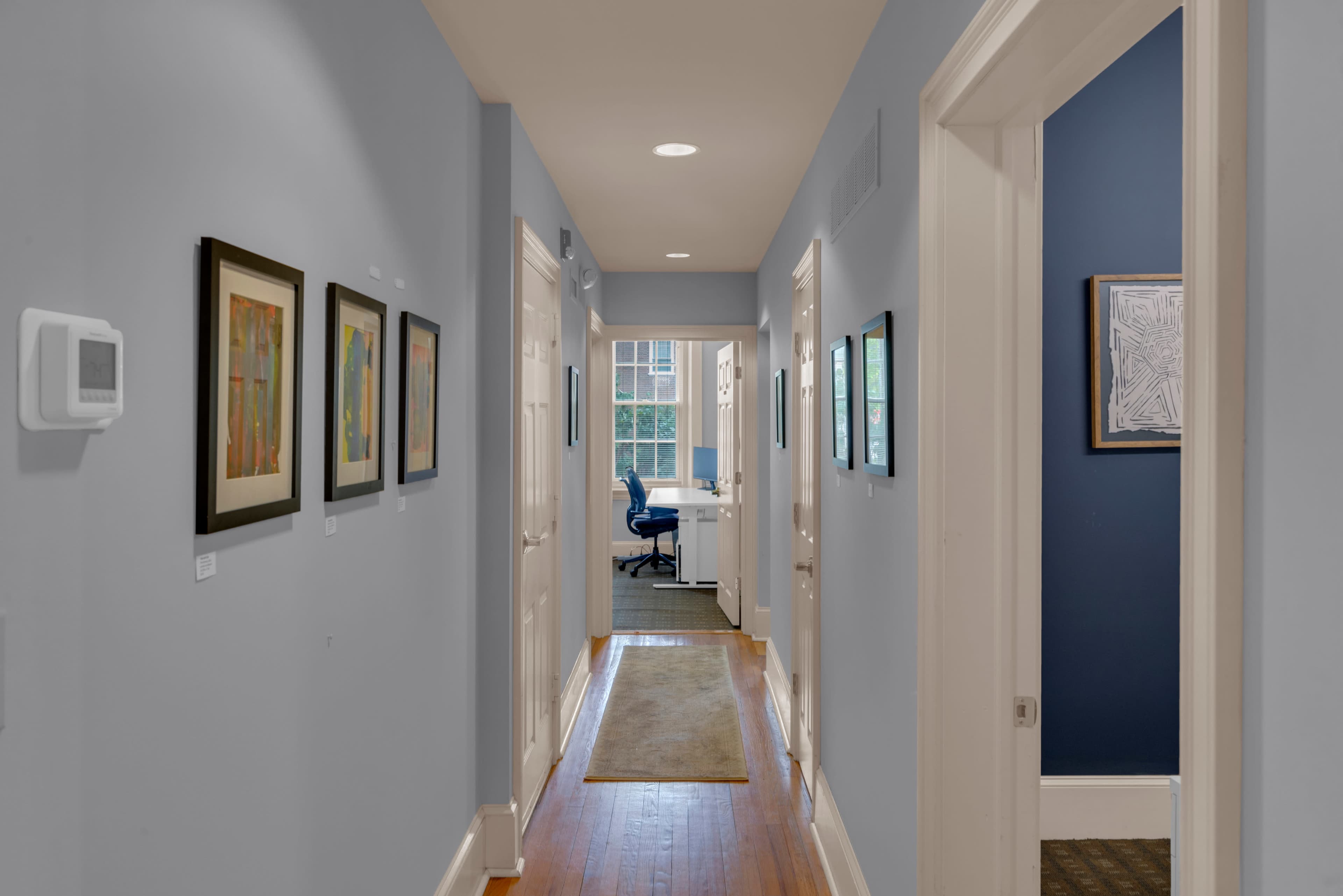 A hallway features walls adorned with framed artwork, leading to a doorway that opens into a room with a desk and chair.