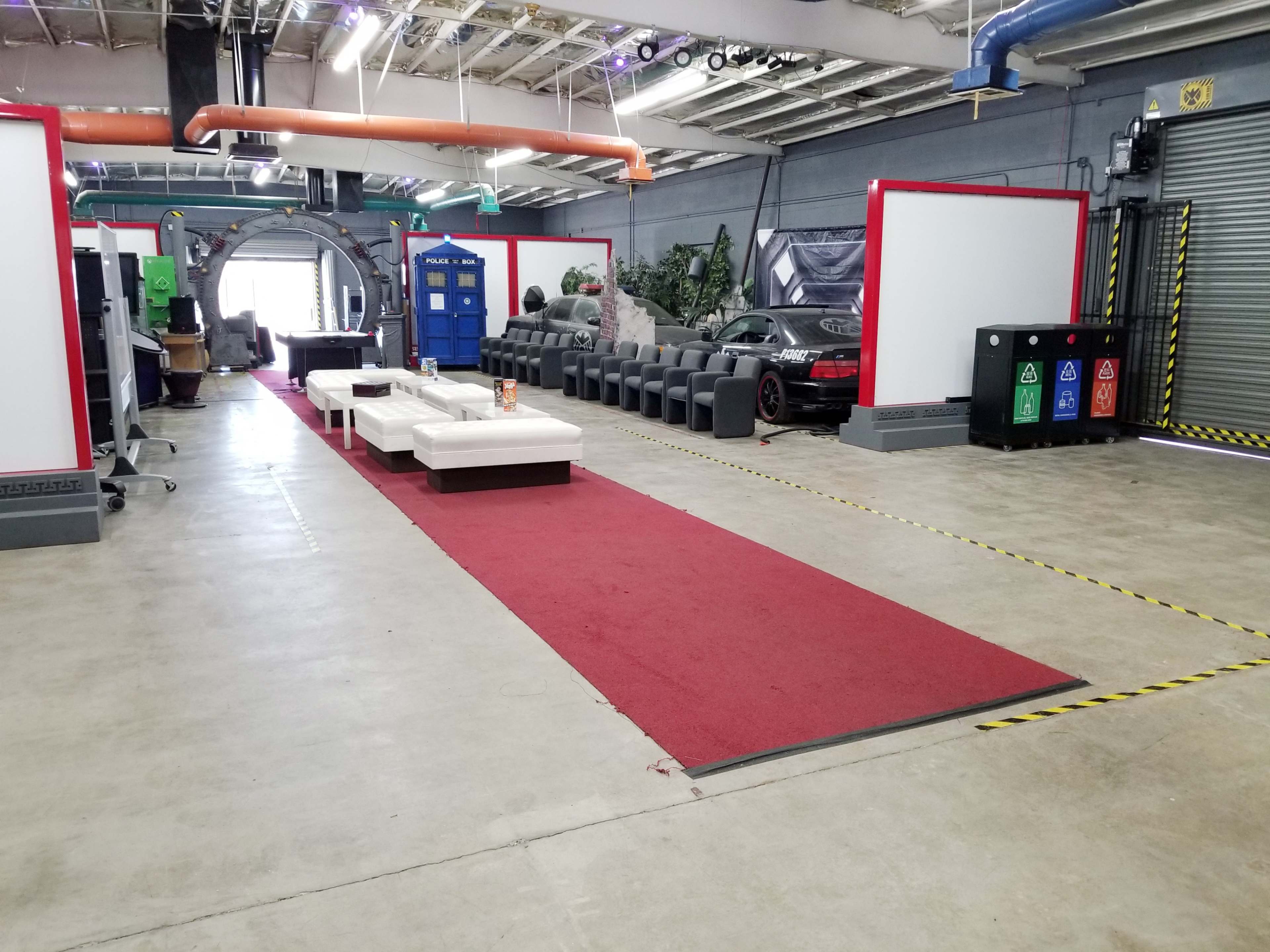 A spacious indoor area with a red carpet leading through rows of seating and a TARDIS, alongside a parked car and recycling bins.