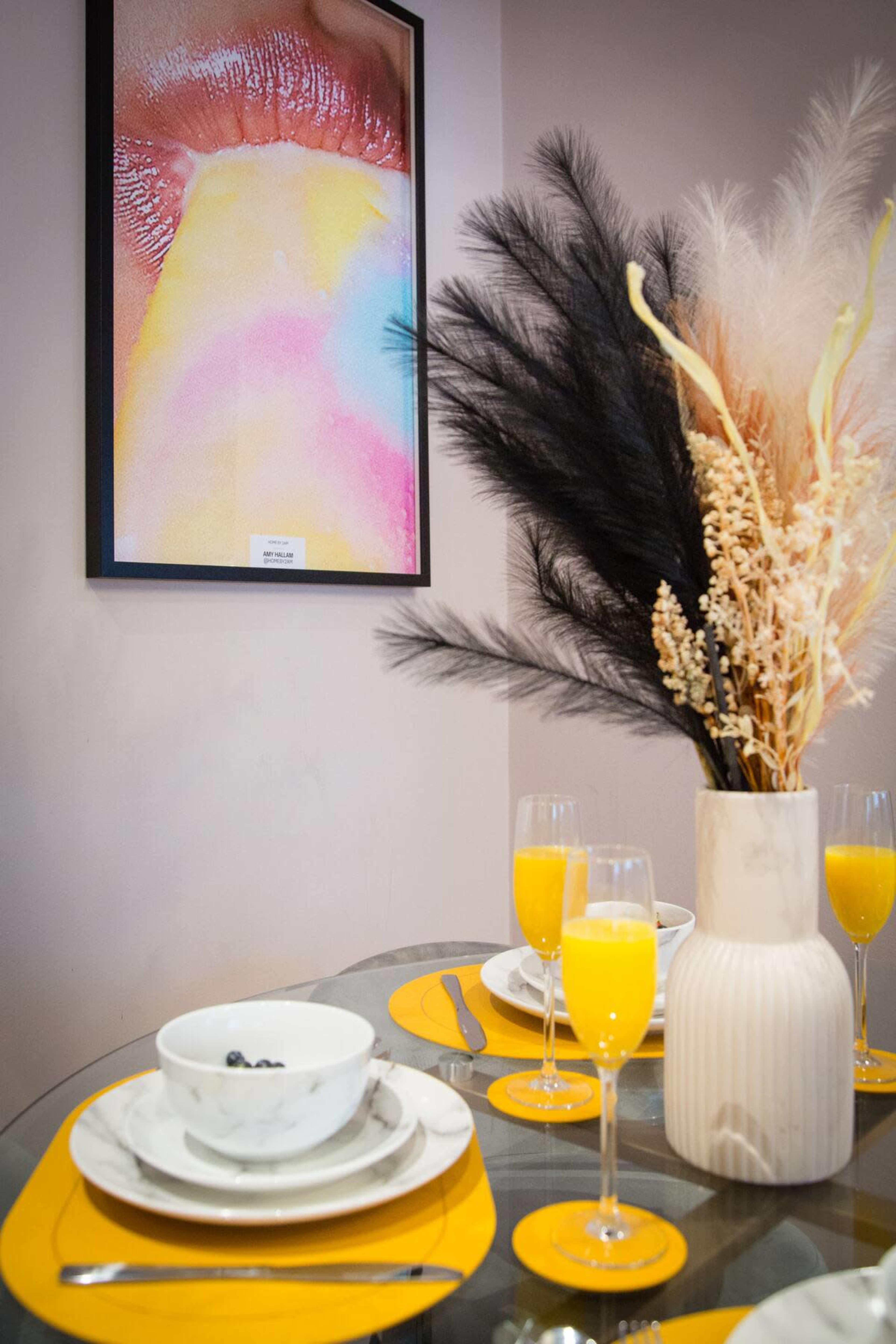 A table set with bowls, glasses of orange juice, and a vase of dried plants is positioned next to a colorful framed artwork of a mouth with a dripping yellow substance.