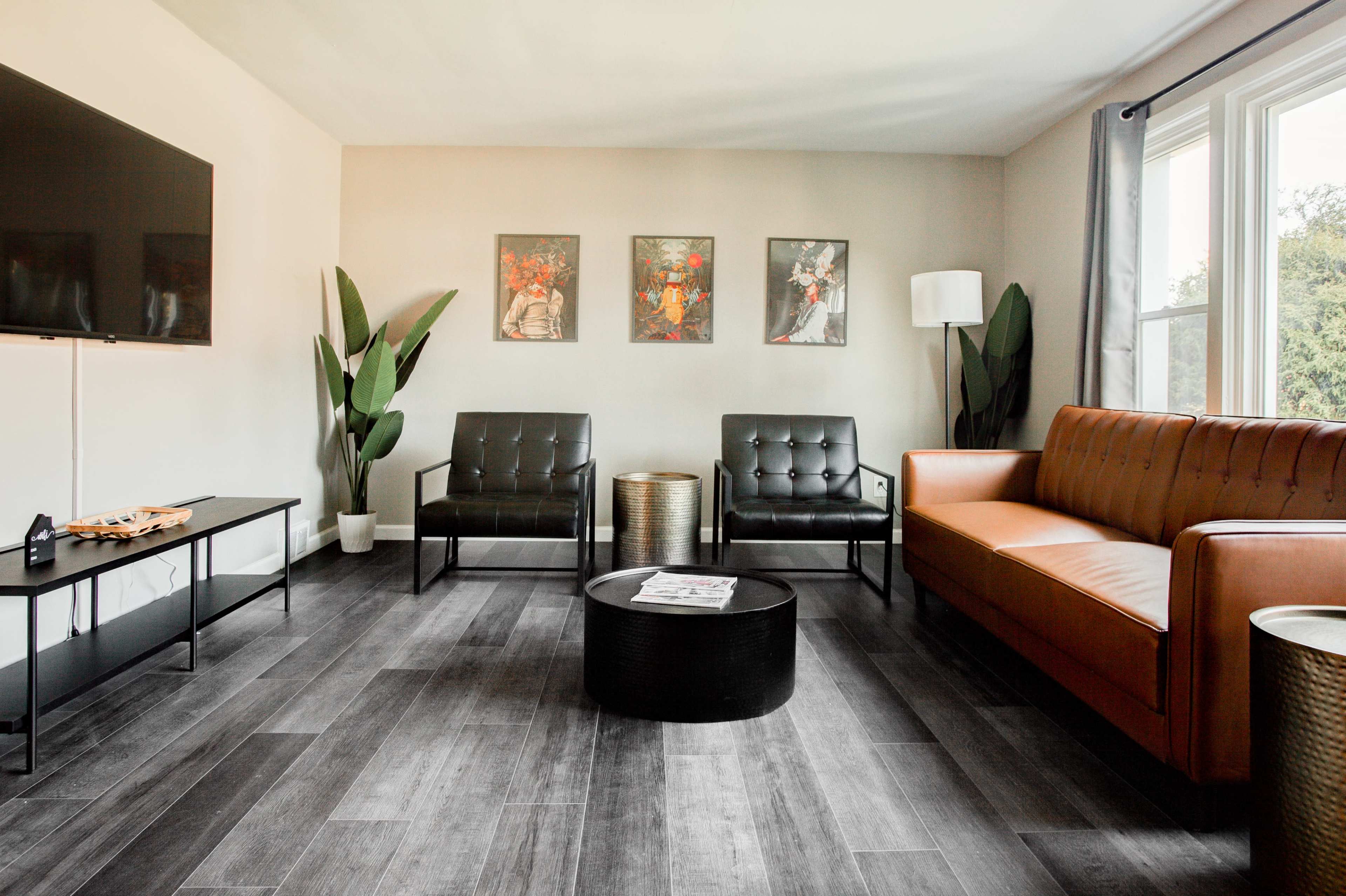 A modern living room features a brown leather sofa, two black armchairs, a round coffee table, and decorative wall art, all set against light-colored walls and wood-style flooring.