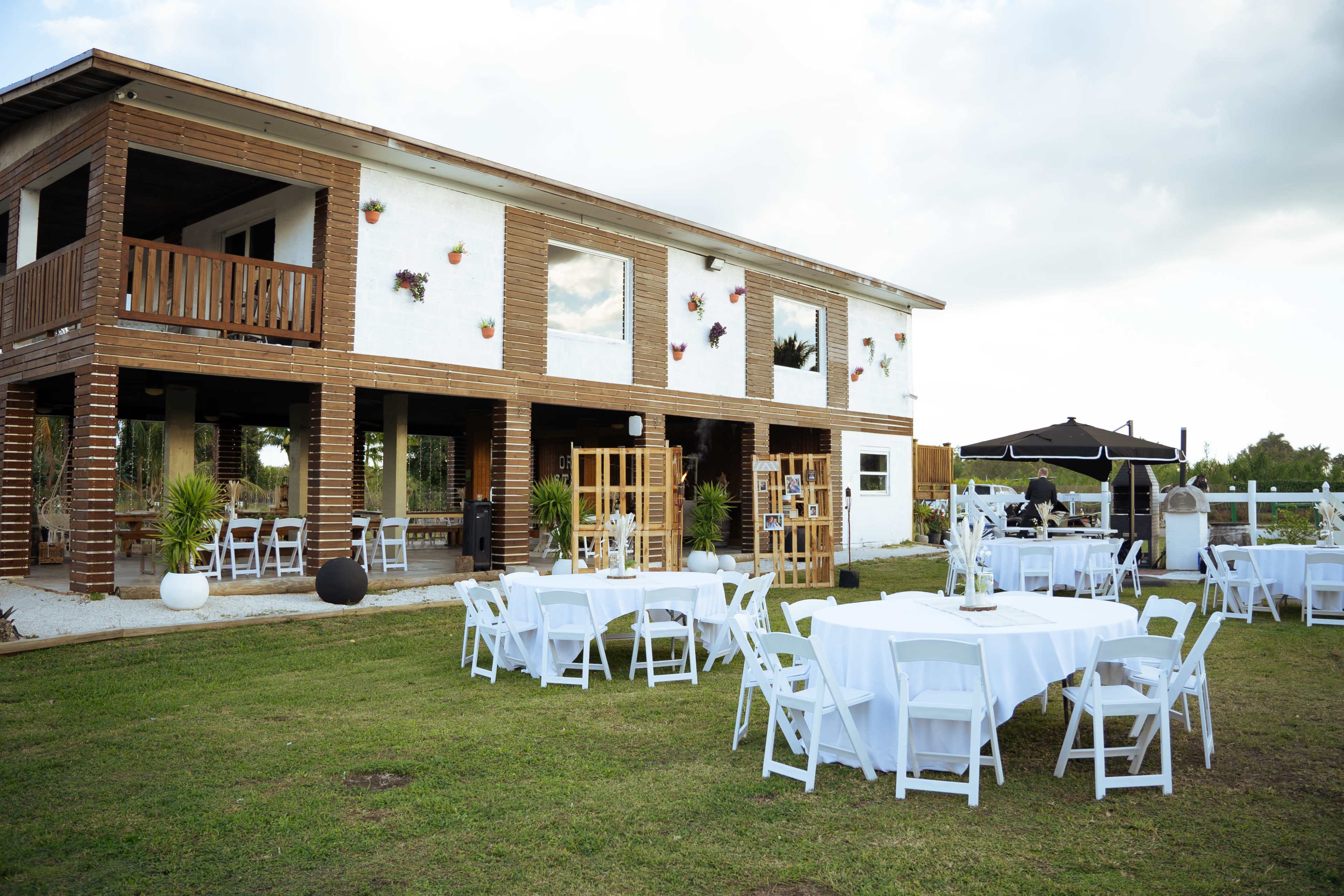 The image displays a two-story building with a rustic facade, surrounded by white tables and chairs set on a grassy area for an outdoor event.