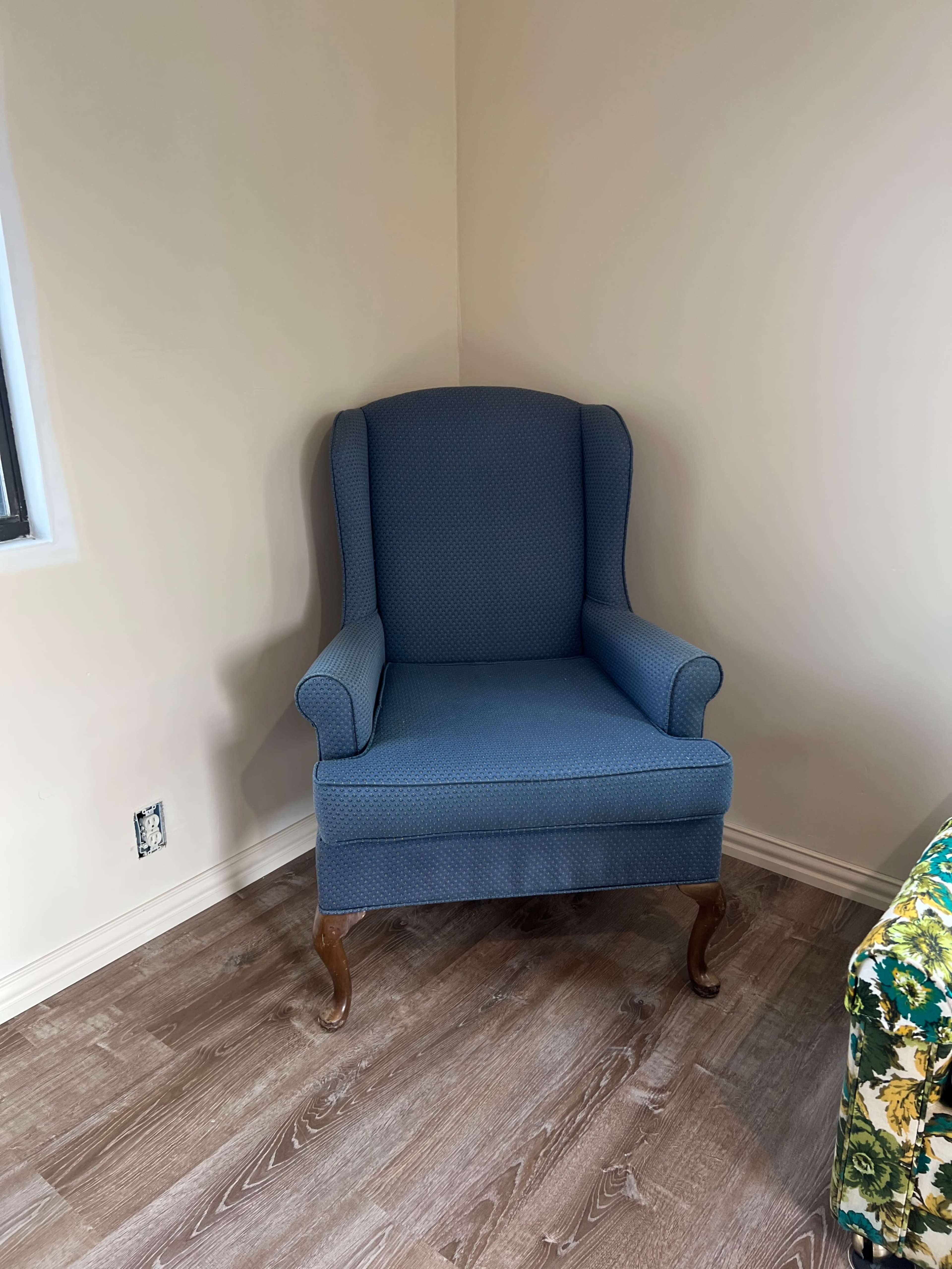 A blue upholstered wingback chair is positioned in the corner of a room with wooden flooring.
