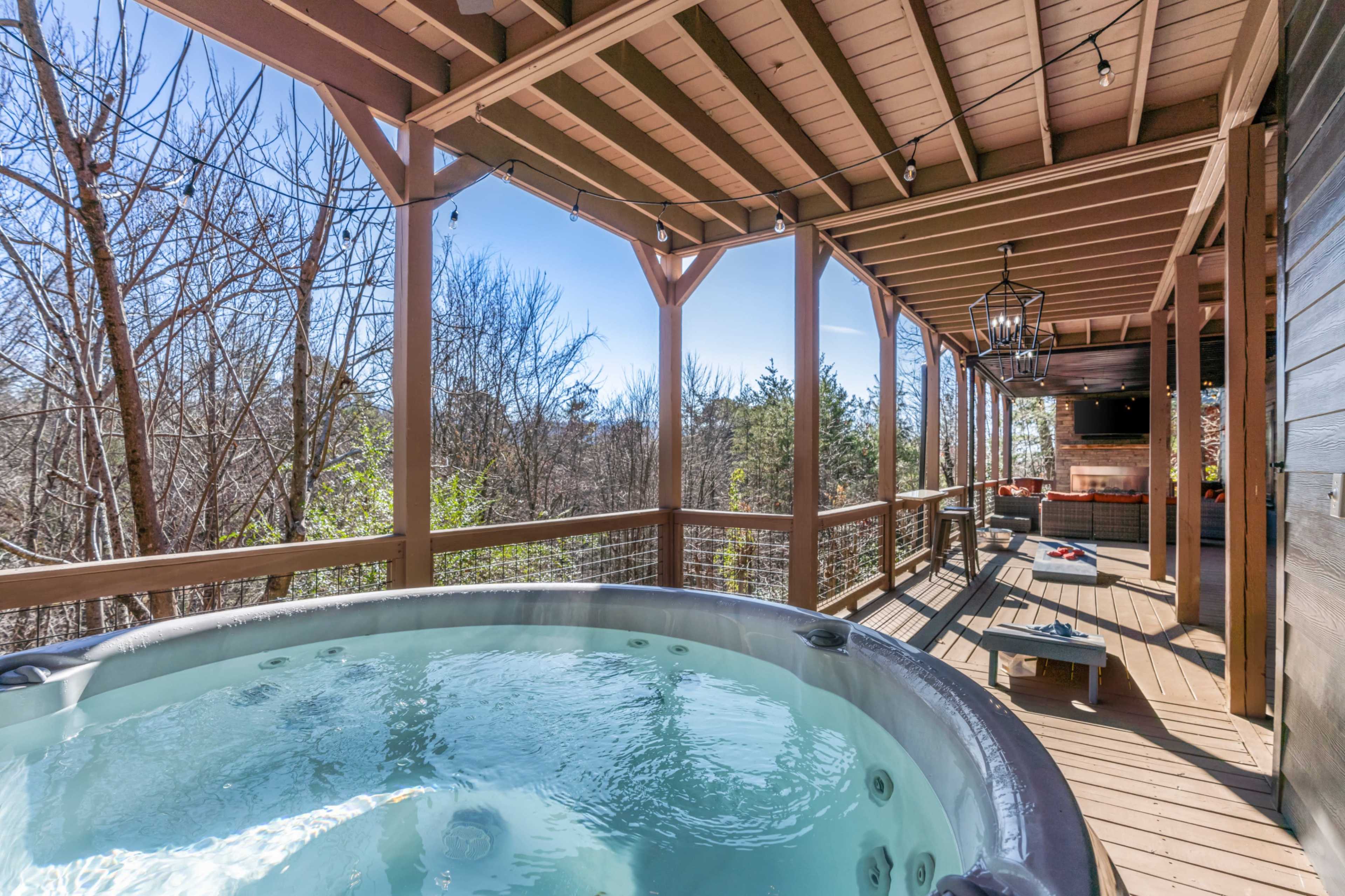 A hot tub sits on a wooden deck under a covered porch, with trees and a clear sky visible in the background.